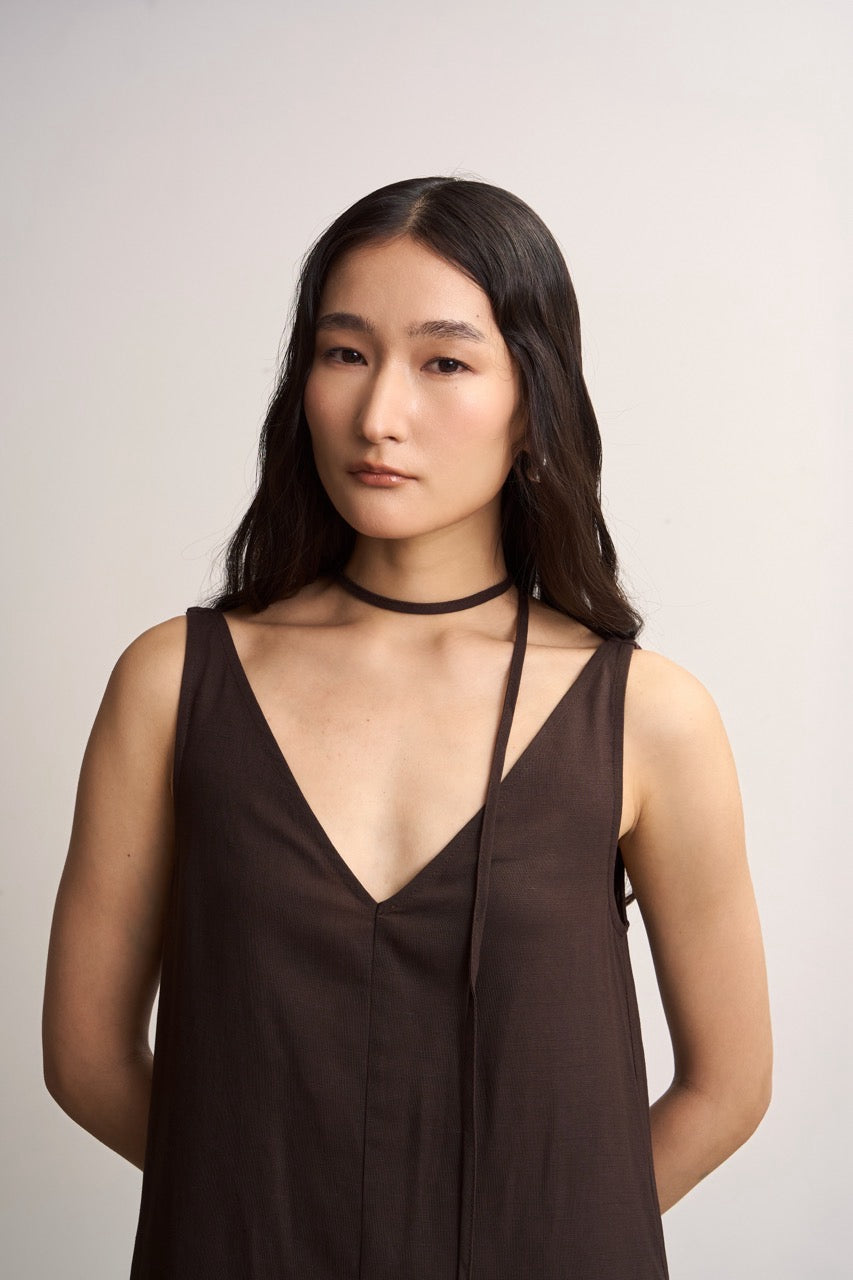 Woman wearing a brown sleeveless linen dress with a tie choker details against a plain background