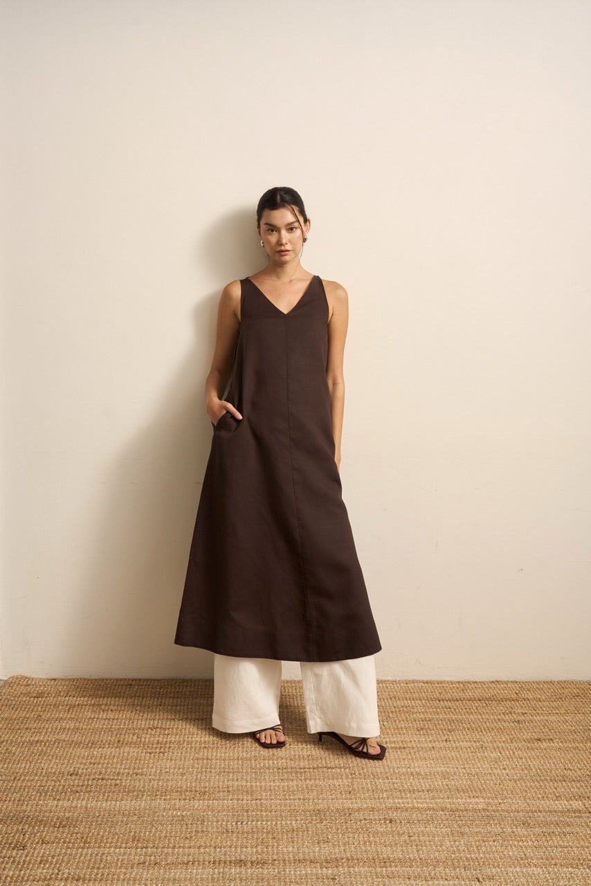 Woman wearing a brown sleeveless linen dress and white linen trousers standing against a beige wall.