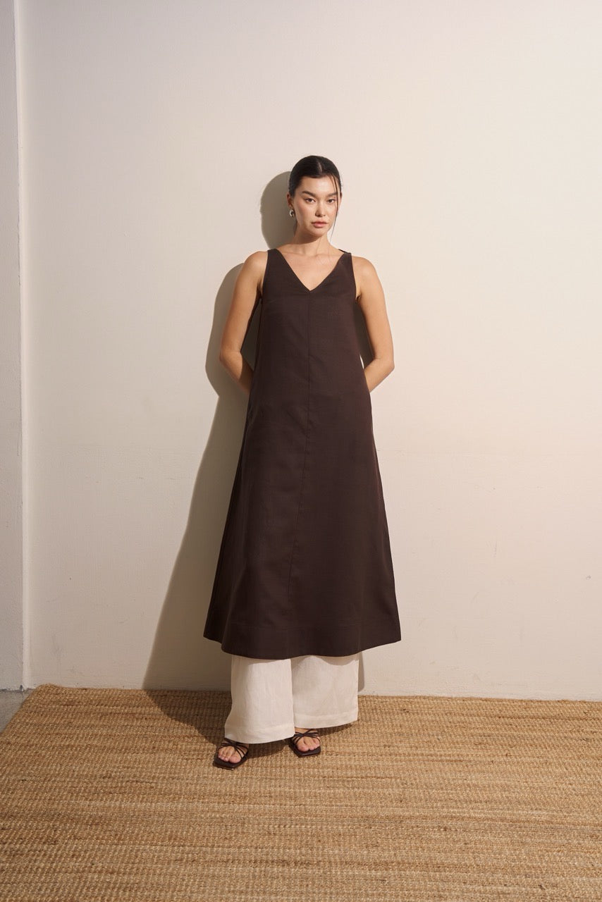 Woman wearing a brown sleeveless linen dress and white linen trousers standing against a plain wall.