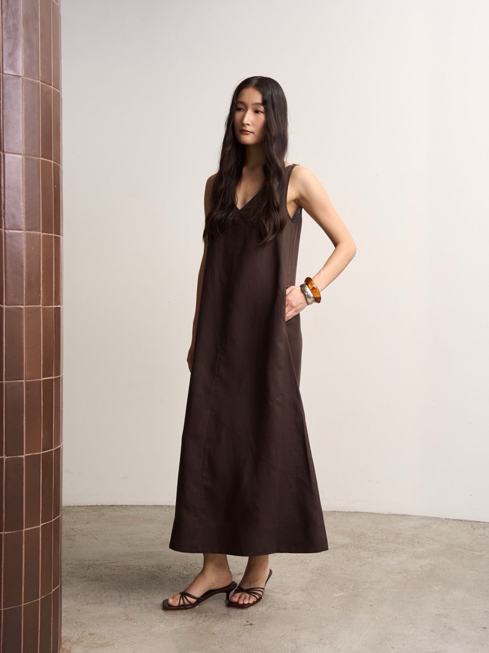 Woman wearing a brown linen dress standing by a plain wall.