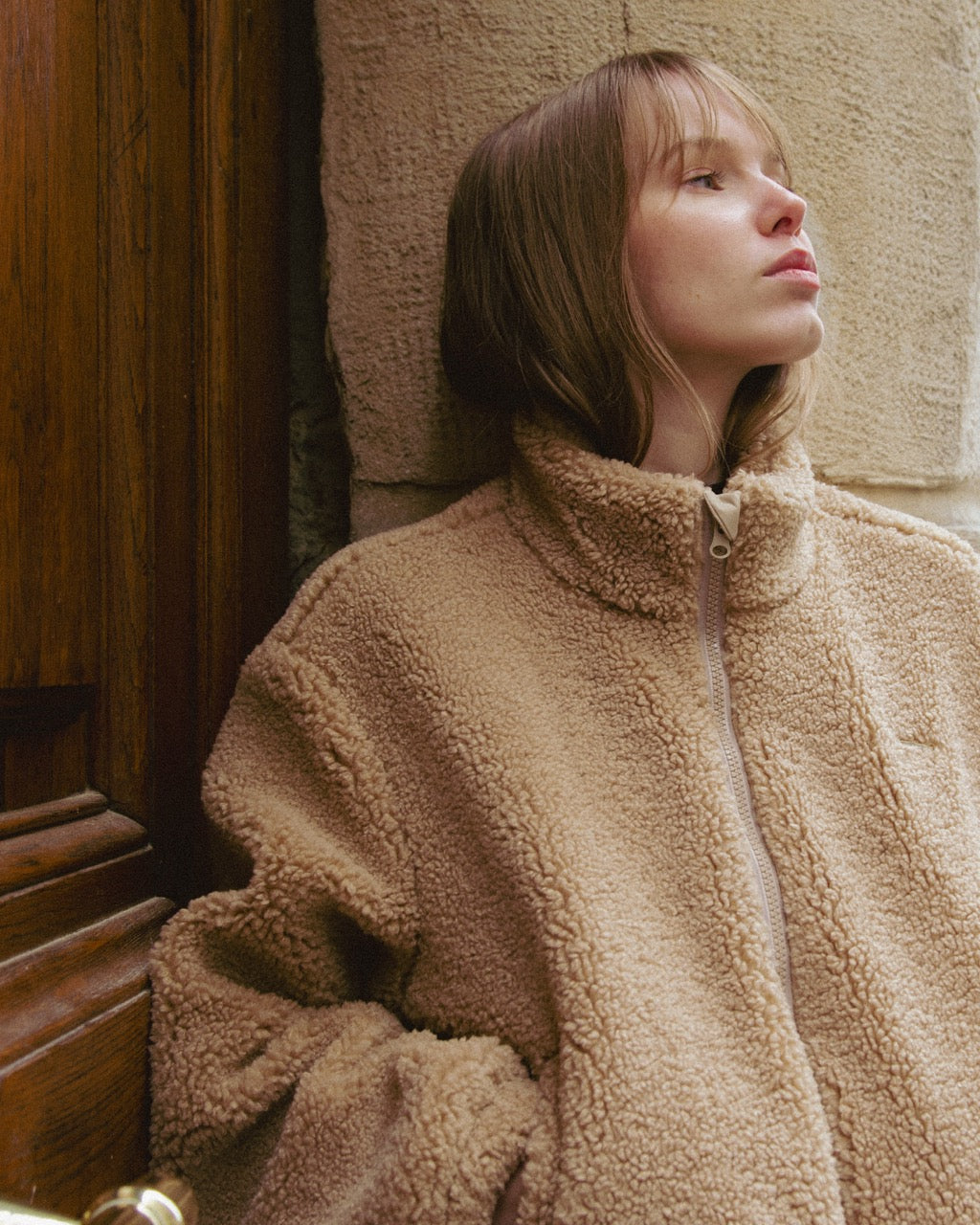woman wearing a beige sherpa coat leaning against a textured wall.