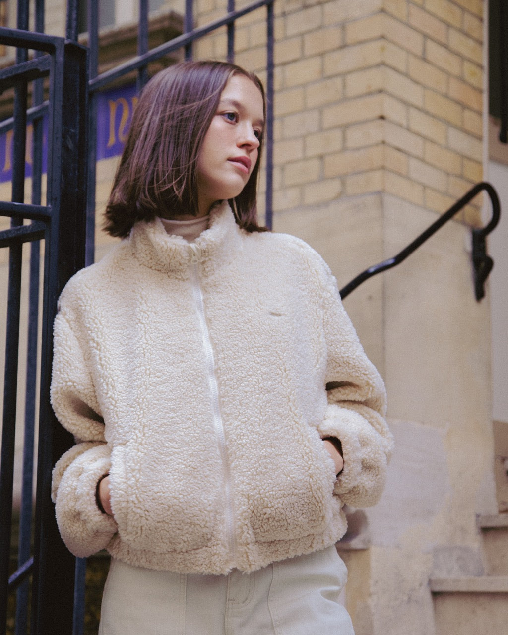 woman wearing a beige fleece jacket standing outdoors near a building.