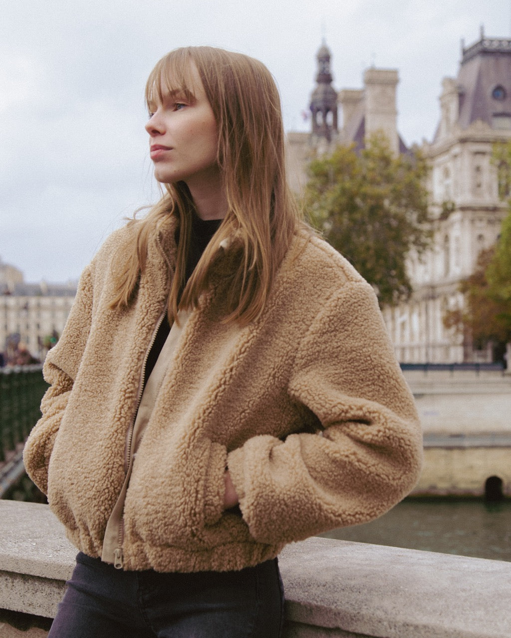 Woman wearing a beige fleece jacket in an urban setting with buildings and water in the background
