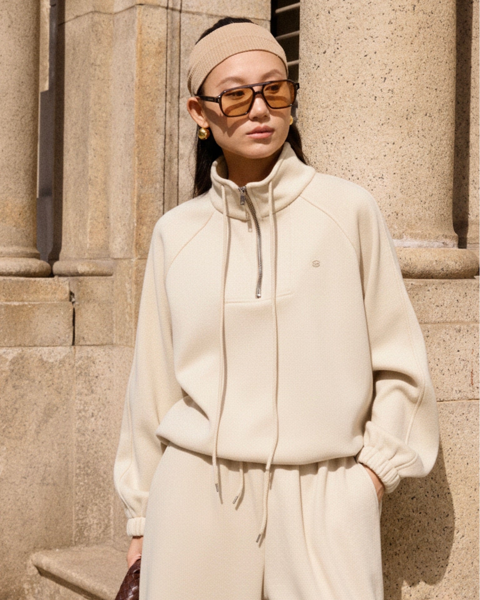 woman wearing a beige tracksuit standing against a stone wall.