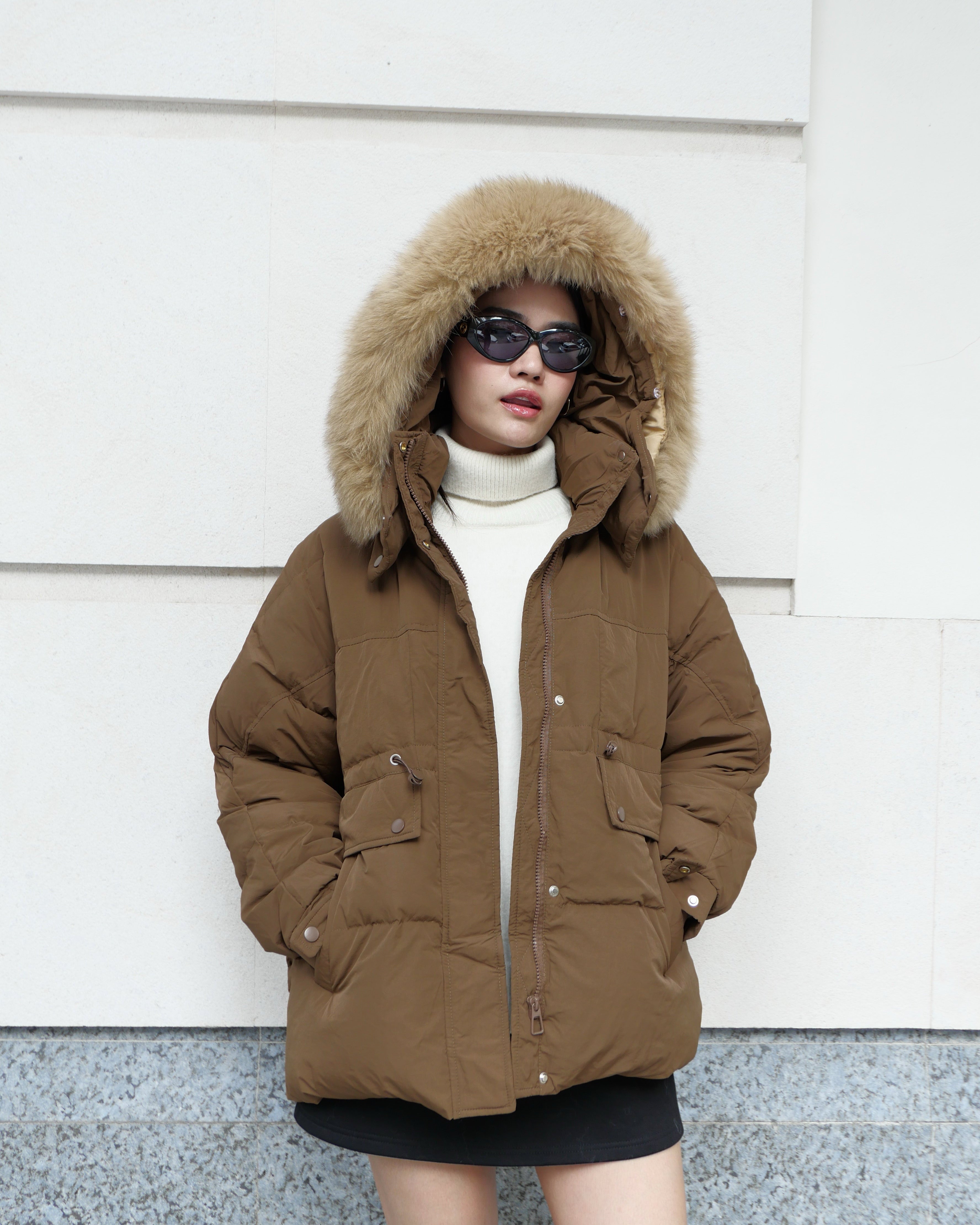 Person wearing a brown winter coat with a fur-lined hood against a white wall.