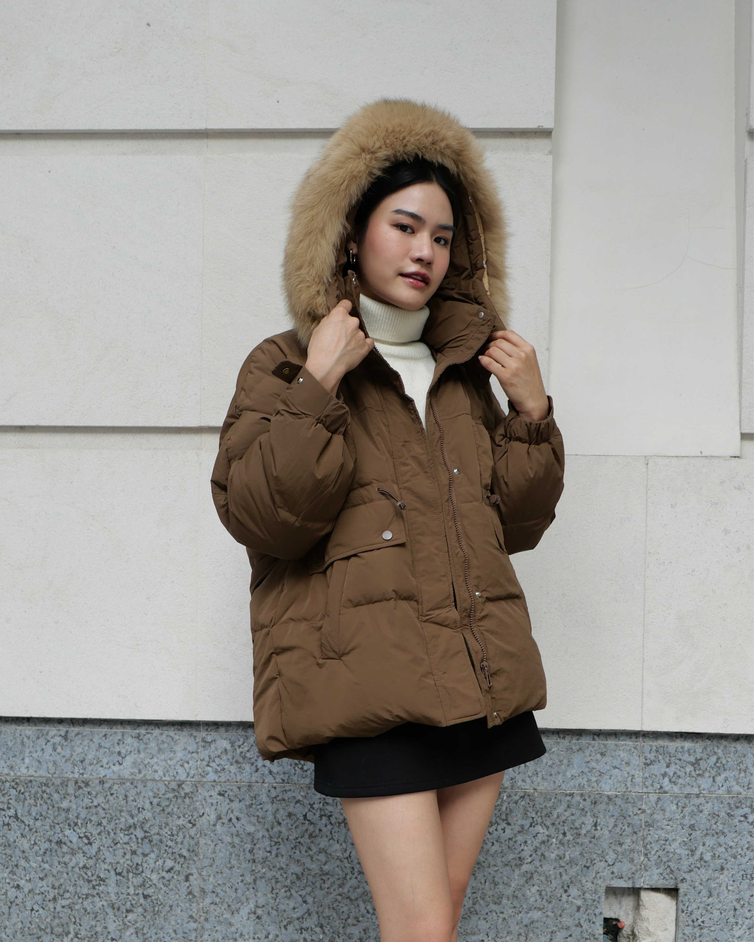 Woman wearing a brown puffer jacket with fur hood against a light-colored wall.