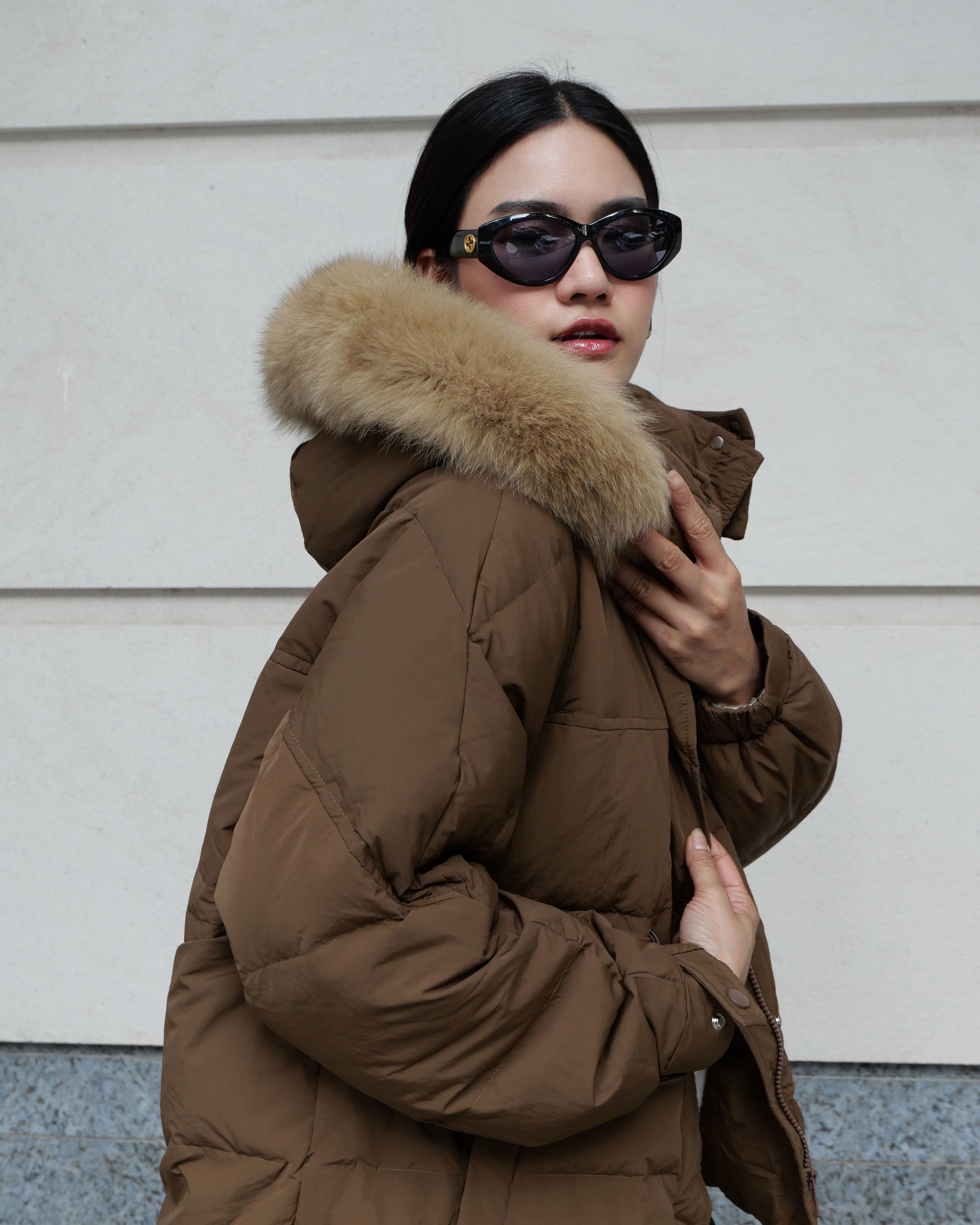 Person wearing a brown puffer jacket with a fur-lined hood against a light-colored wall.