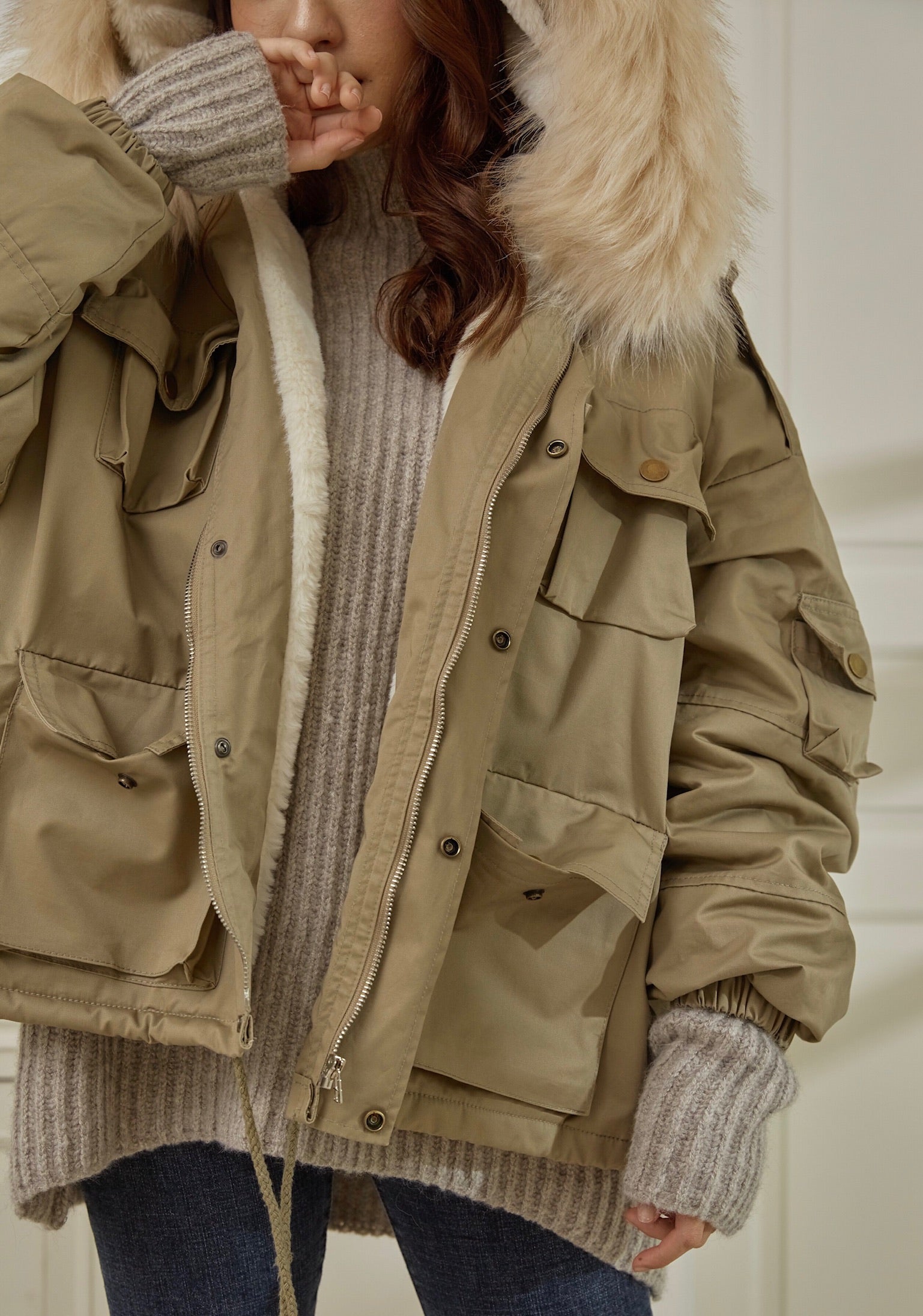 woman wearing a beige parka with fur hood, gray sweater, and gloves indoors.