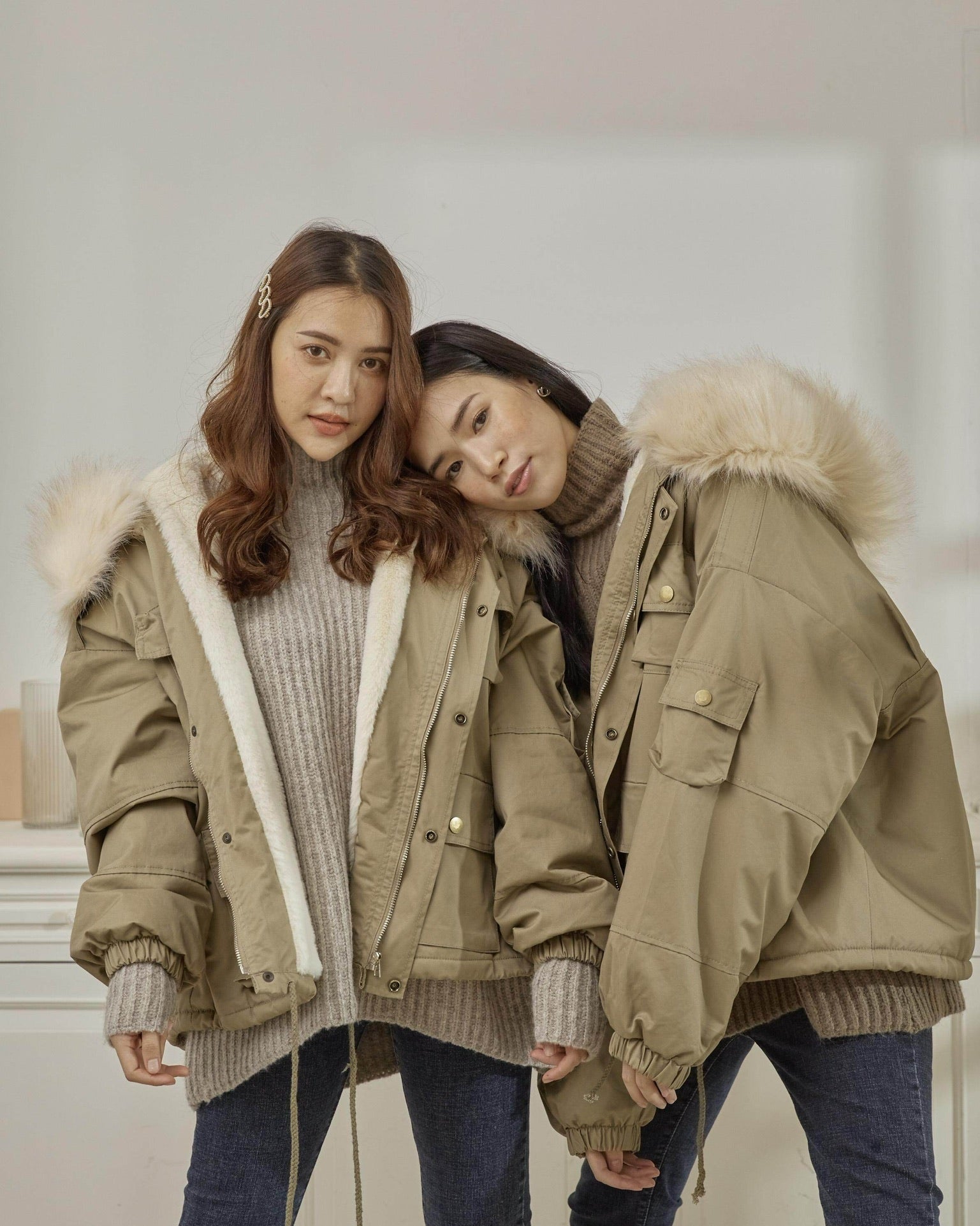 Two women wearing matching beige parkas with fur hoods indoors.