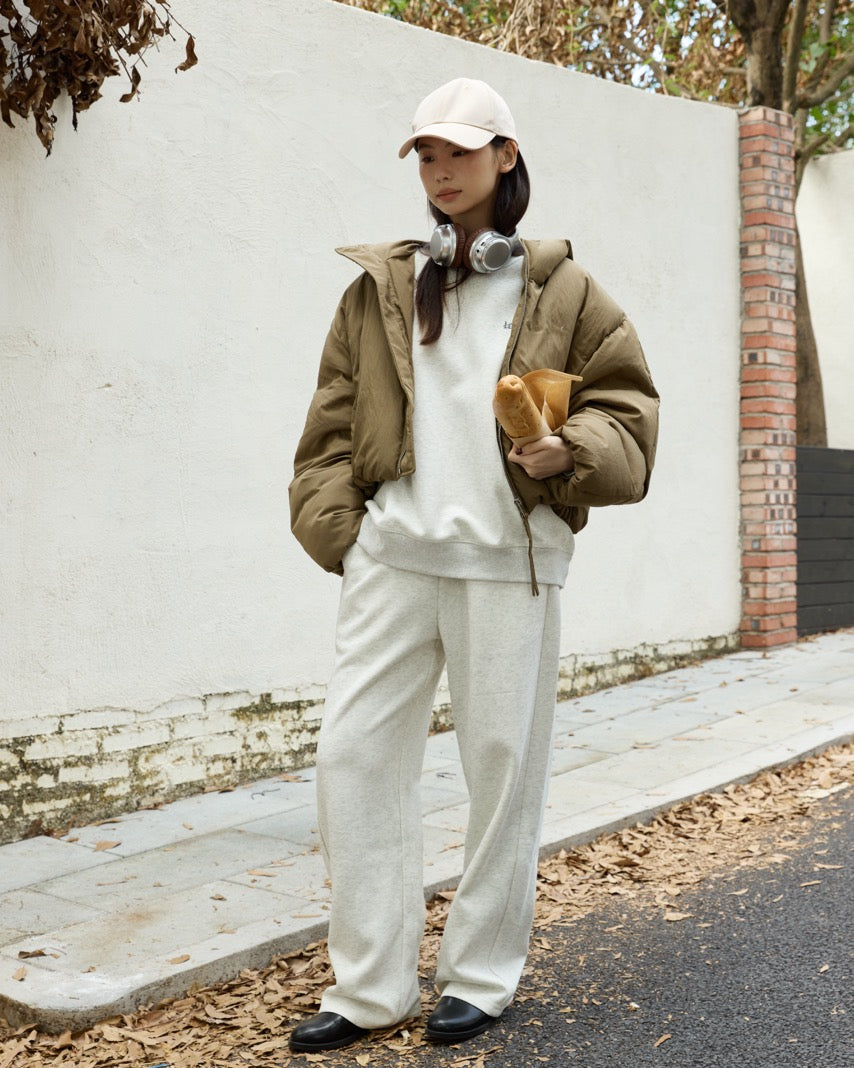 woman wearing a green jacket, white sweater, and pants, holding a brown bag, standing against a white wall.