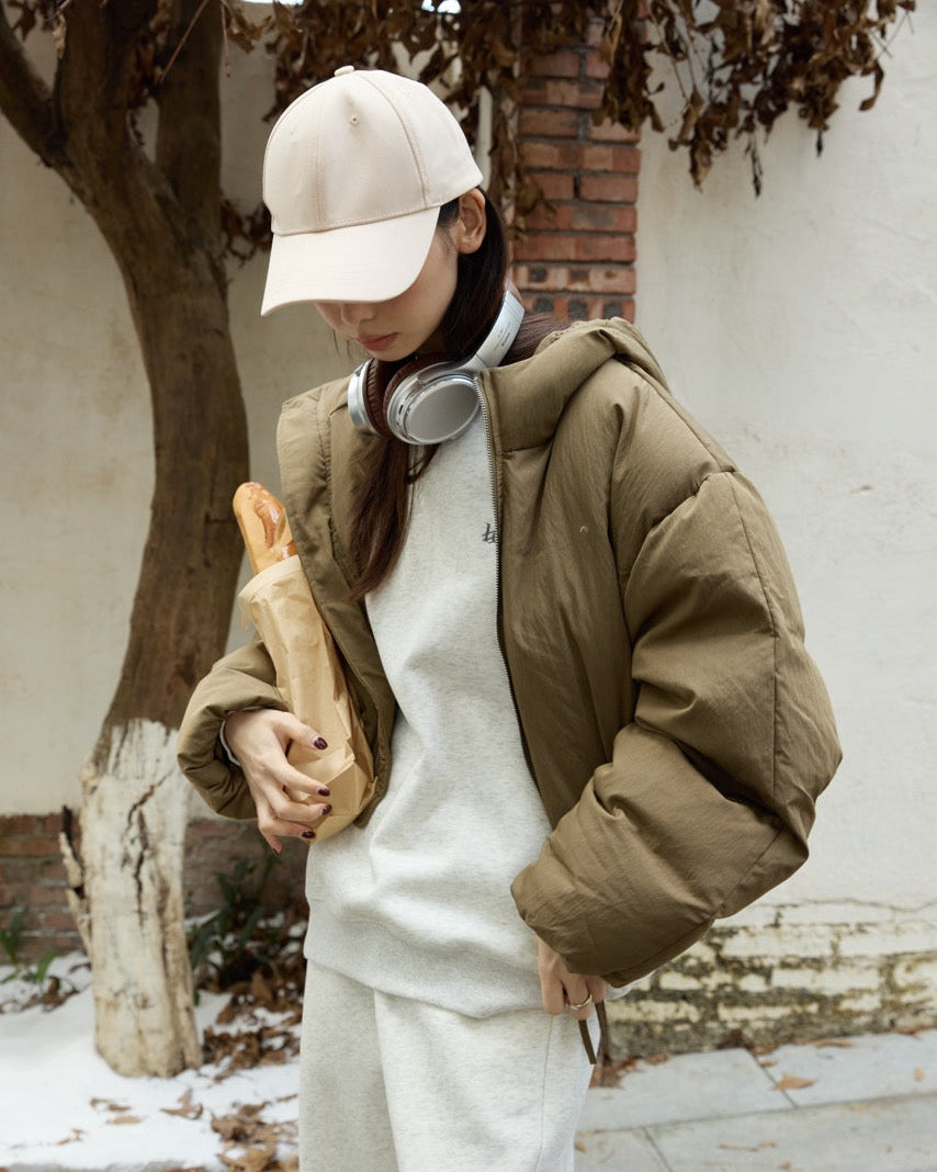 woman wearing a beige cap, green puffer jacket, and white outfit holding bread outdoors.