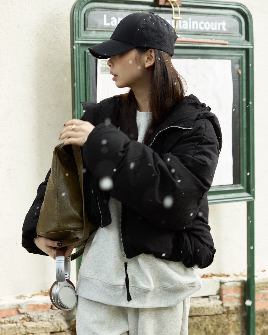 woman wearing a black jacket and cap, holding a green bag, standing near a green phone booth.