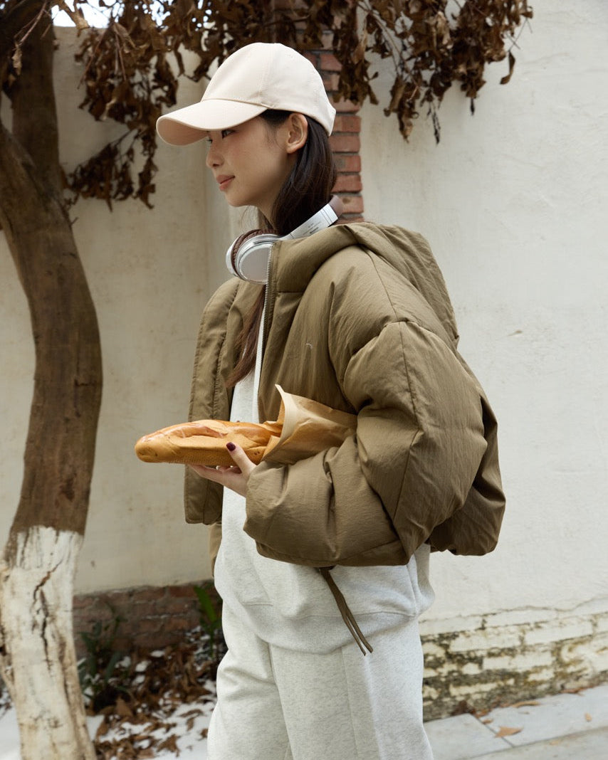 woman wearing a brown puffer jacket and white pants, holding bread outdoors.