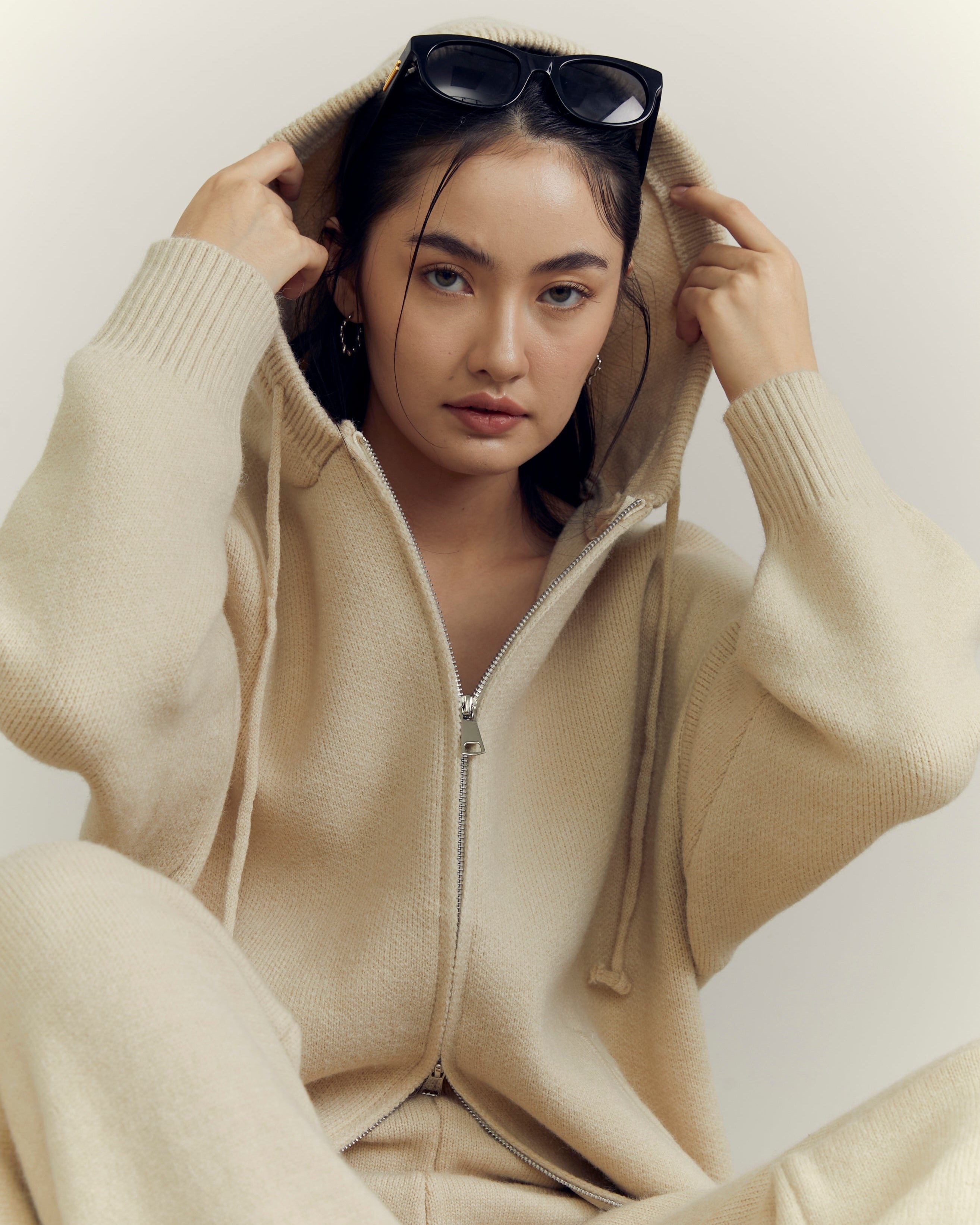 woman wearing a beige hoodie and pants with sunglasses on a plain background