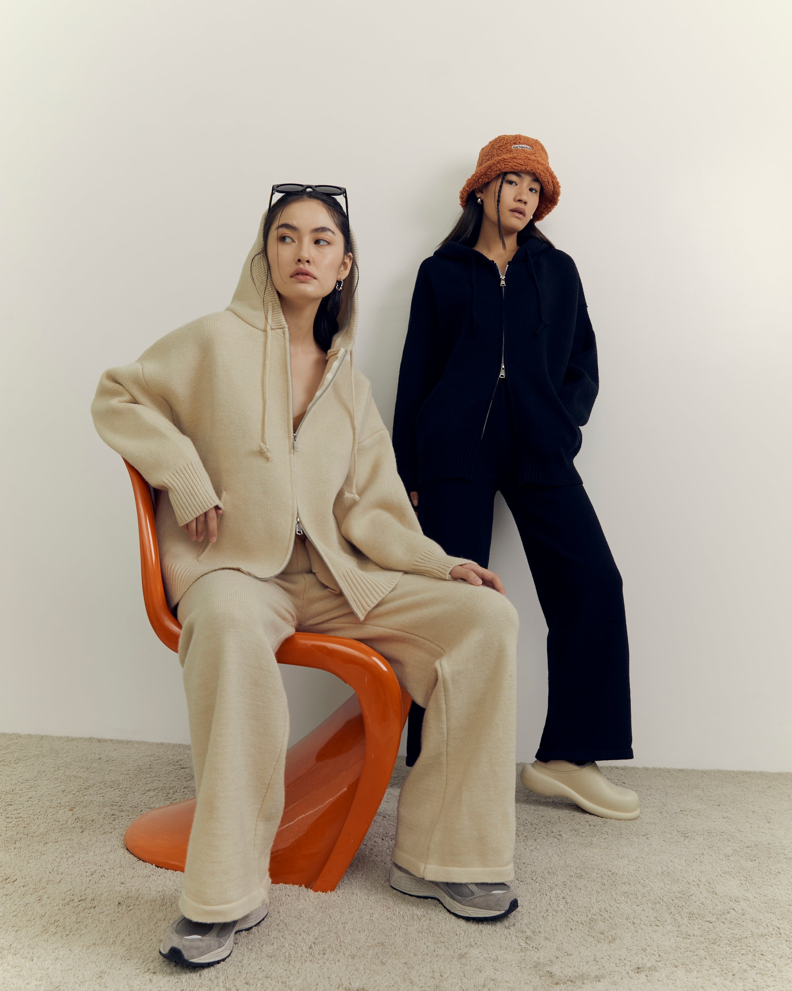 Two women sitting on a white background, one in a beige outfit and the other in black.