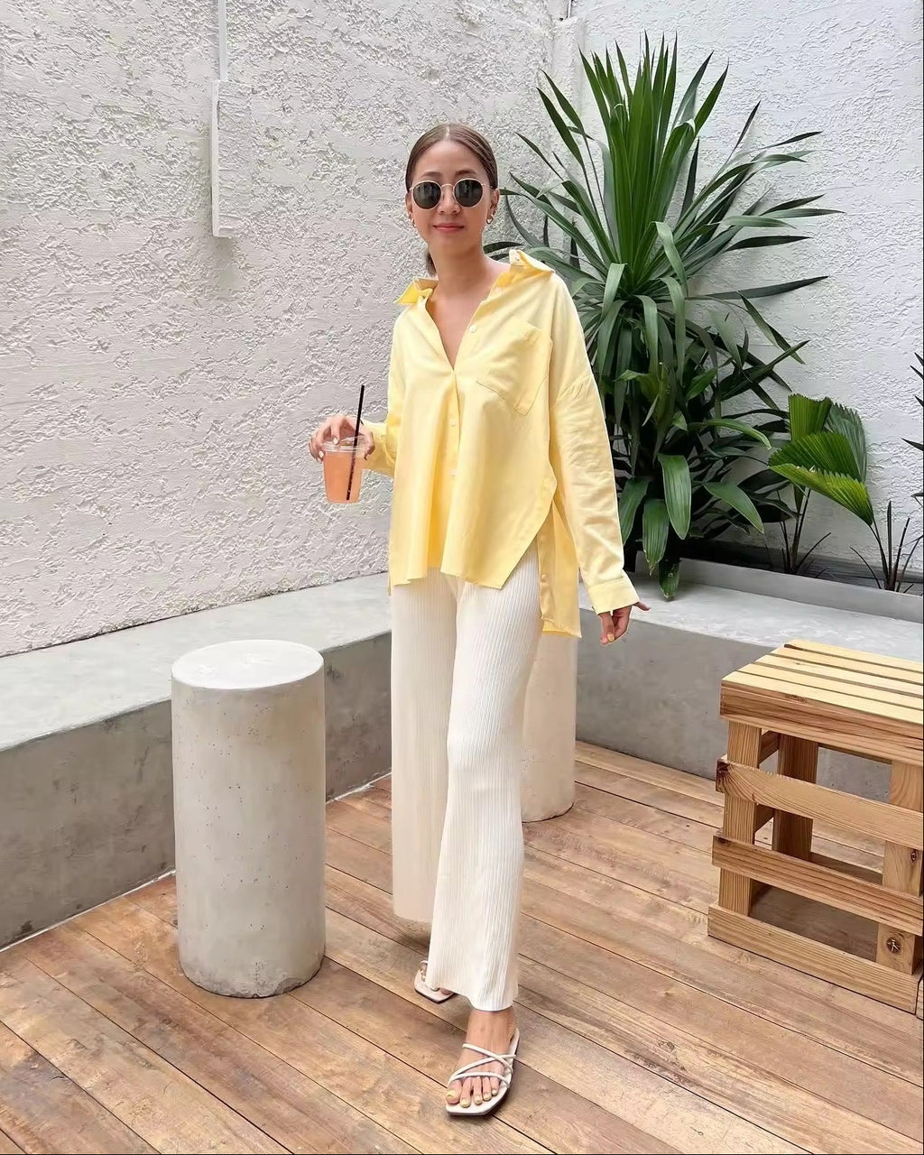 Woman in yellow jacket and white pants standing on a wooden deck with plants and a white wall in the background.