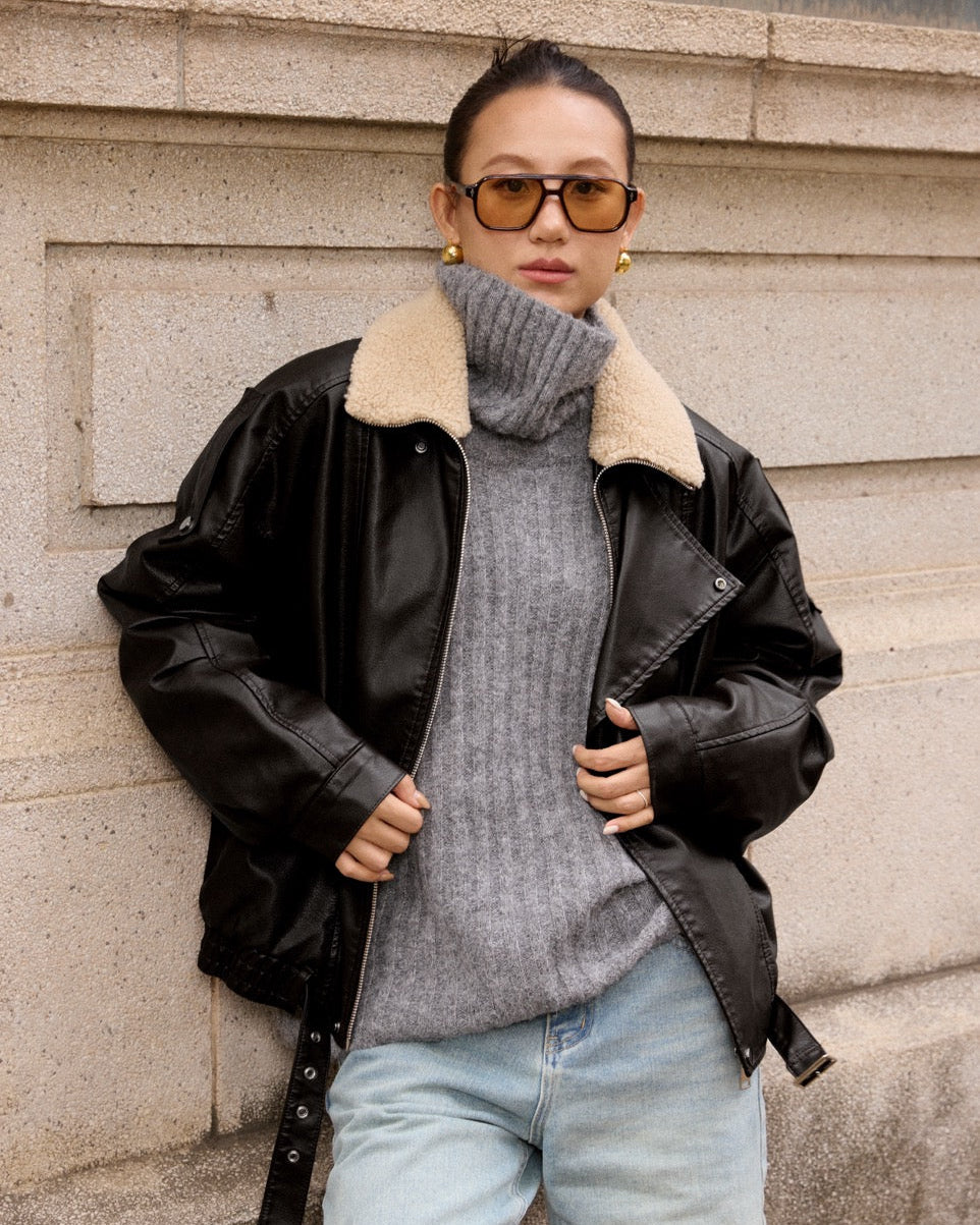 woman wearing a black leather jacket with a beige fur-lined hood, gray sweater, and light blue jeans against a stone wall.