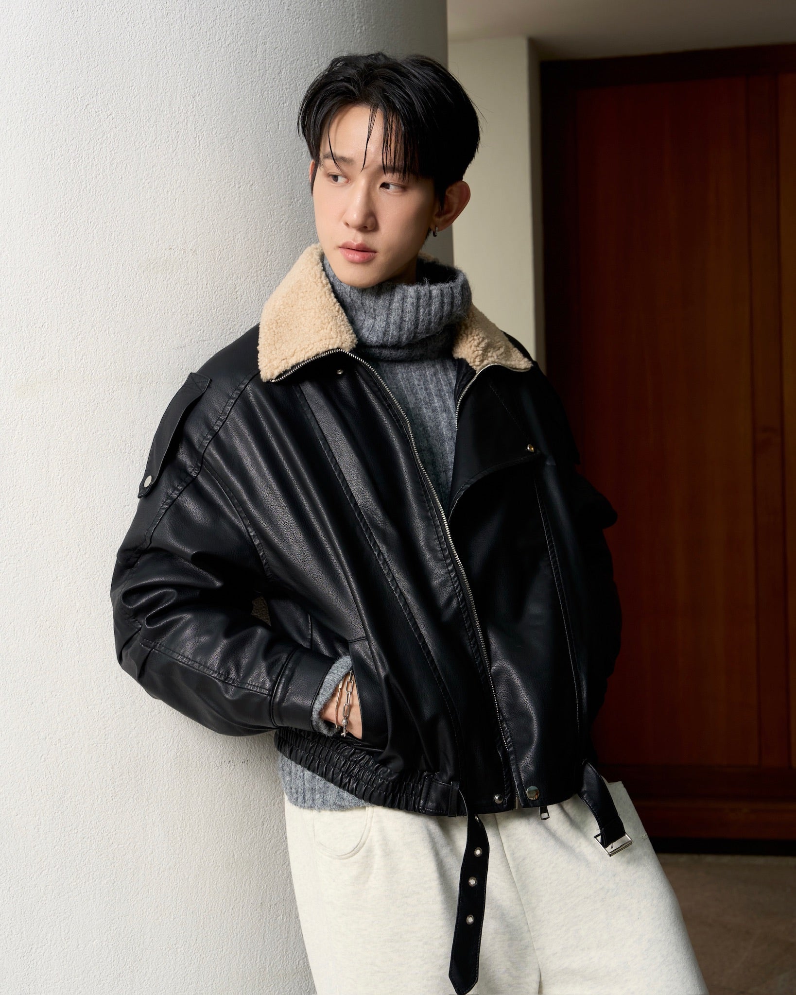 man wearing a black leather jacket with a beige interior, standing against a white wall.