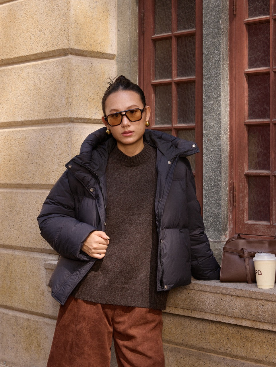 woman wearing a black puffer jacket and sunglasses, standing against a building with a window.