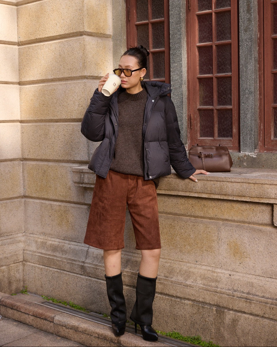 woman standing on steps outside a building, wearing a dark jacket, brown shorts, and black boots, holding a coffee cup.
