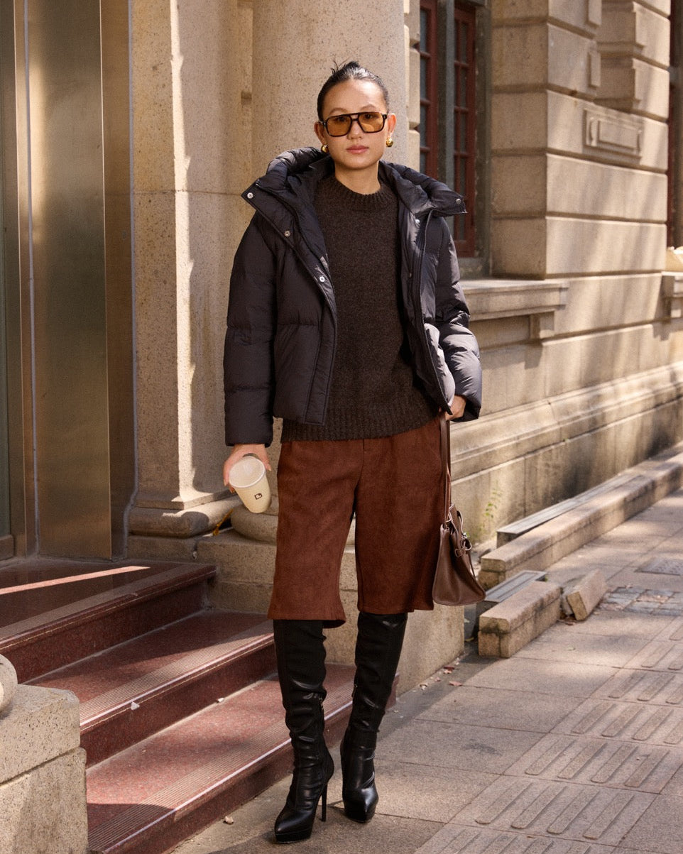 woman wearing a black coat, brown pants, and black boots standing on steps outside a building.