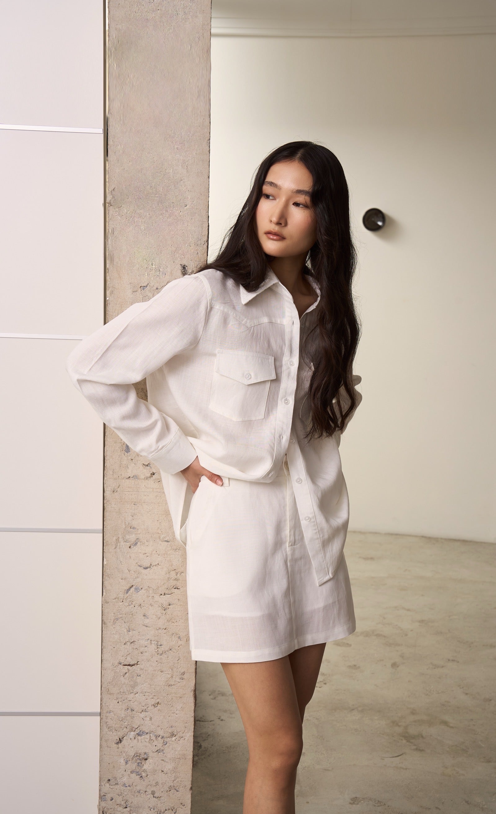 Woman wearing a white linen double pocket shirt with white linen skirt standing against a neutral wall.