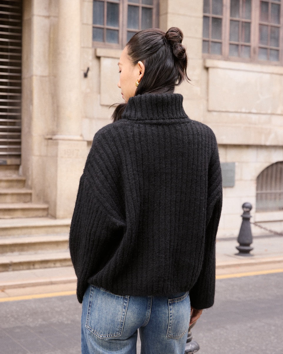 woman wearing a black turtleneck sweater and blue jeans standing in front of a building.