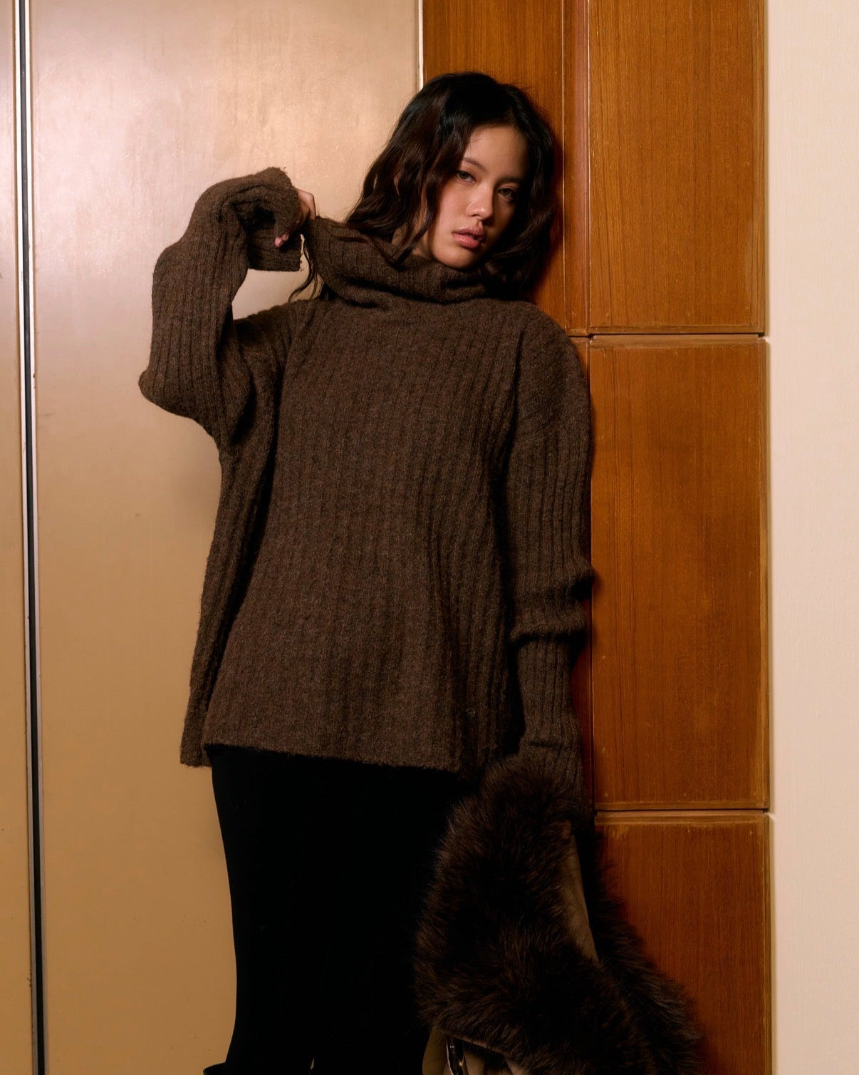 woman wearing a brown sweater and black pants standing against a wooden panel wall.