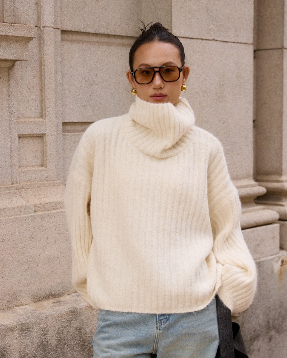 woman wearing a cream turtleneck sweater and light blue jeans standing against a stone wall.