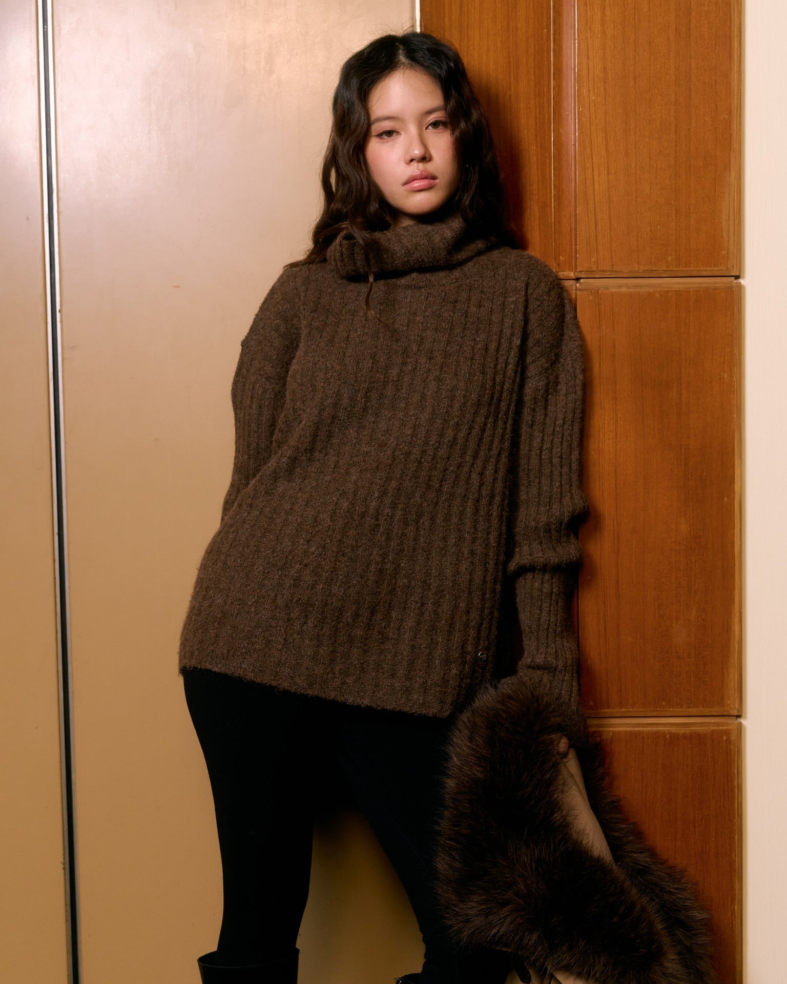 woman wearing a brown sweater and black pants against a wooden panel wall.