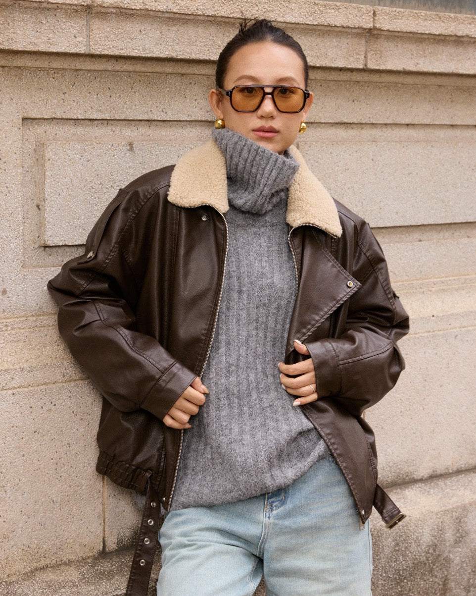 woman wearing a brown leather jacket with a fur-lined hood, gray sweater, and light blue jeans against a stone wall.