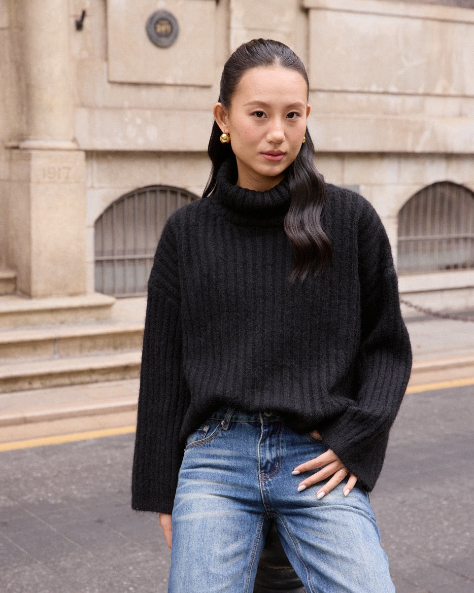 Woman wearing a black sweater and blue jeans standing in front of a building.