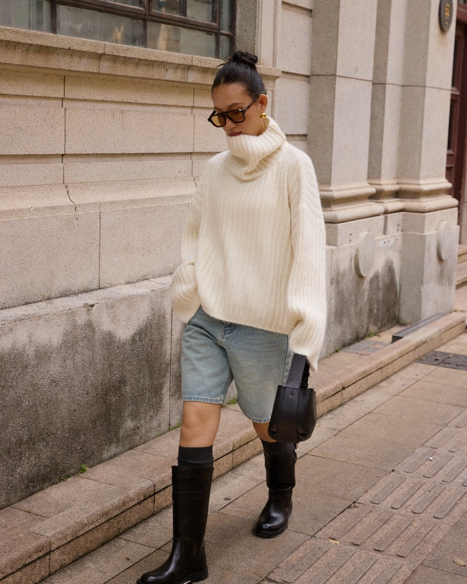 Woman wearing a white sweater, denim shorts, and black boots standing on a street.