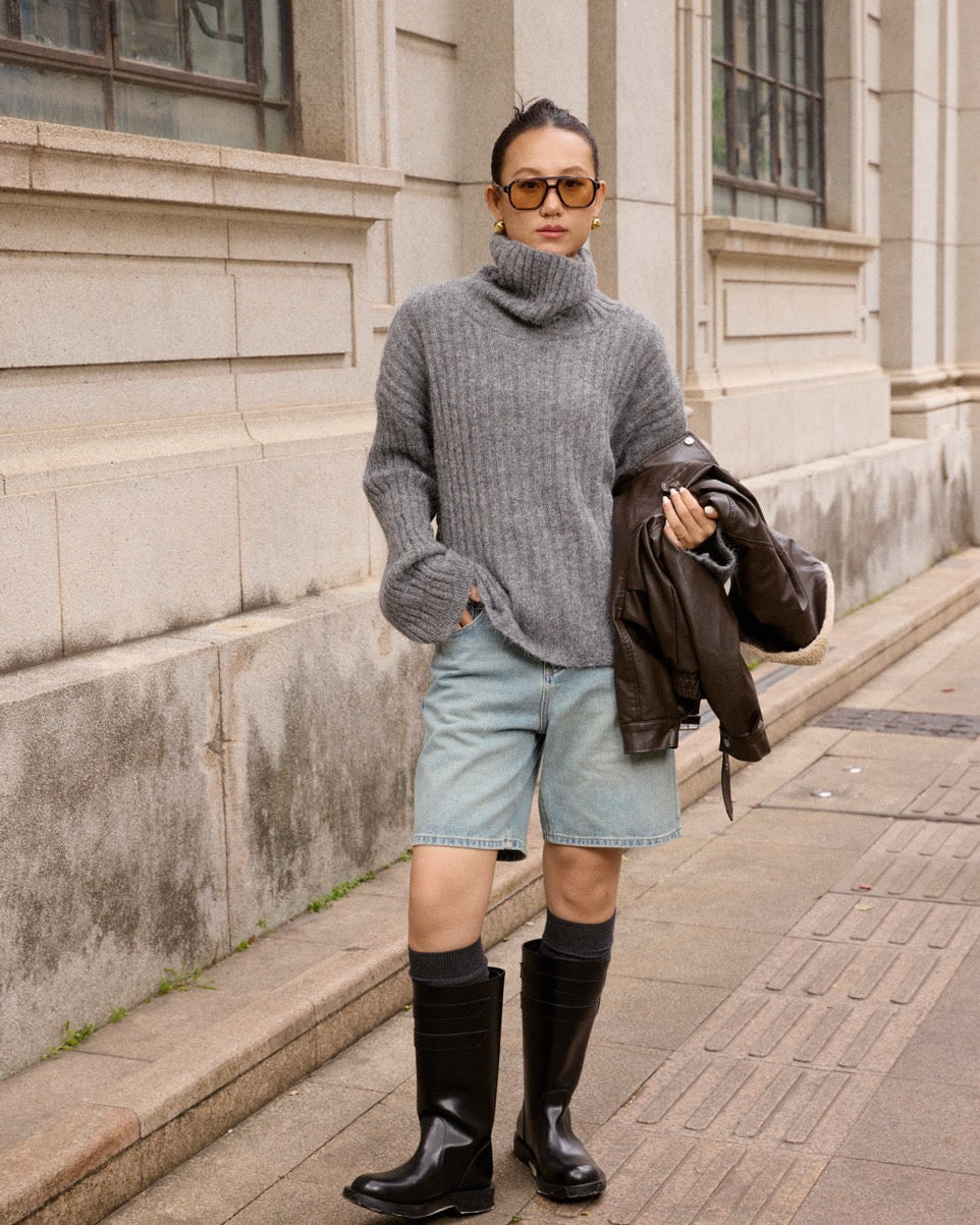 woman wearing a gray sweater, light blue shorts, and black rain boots standing on a sidewalk.