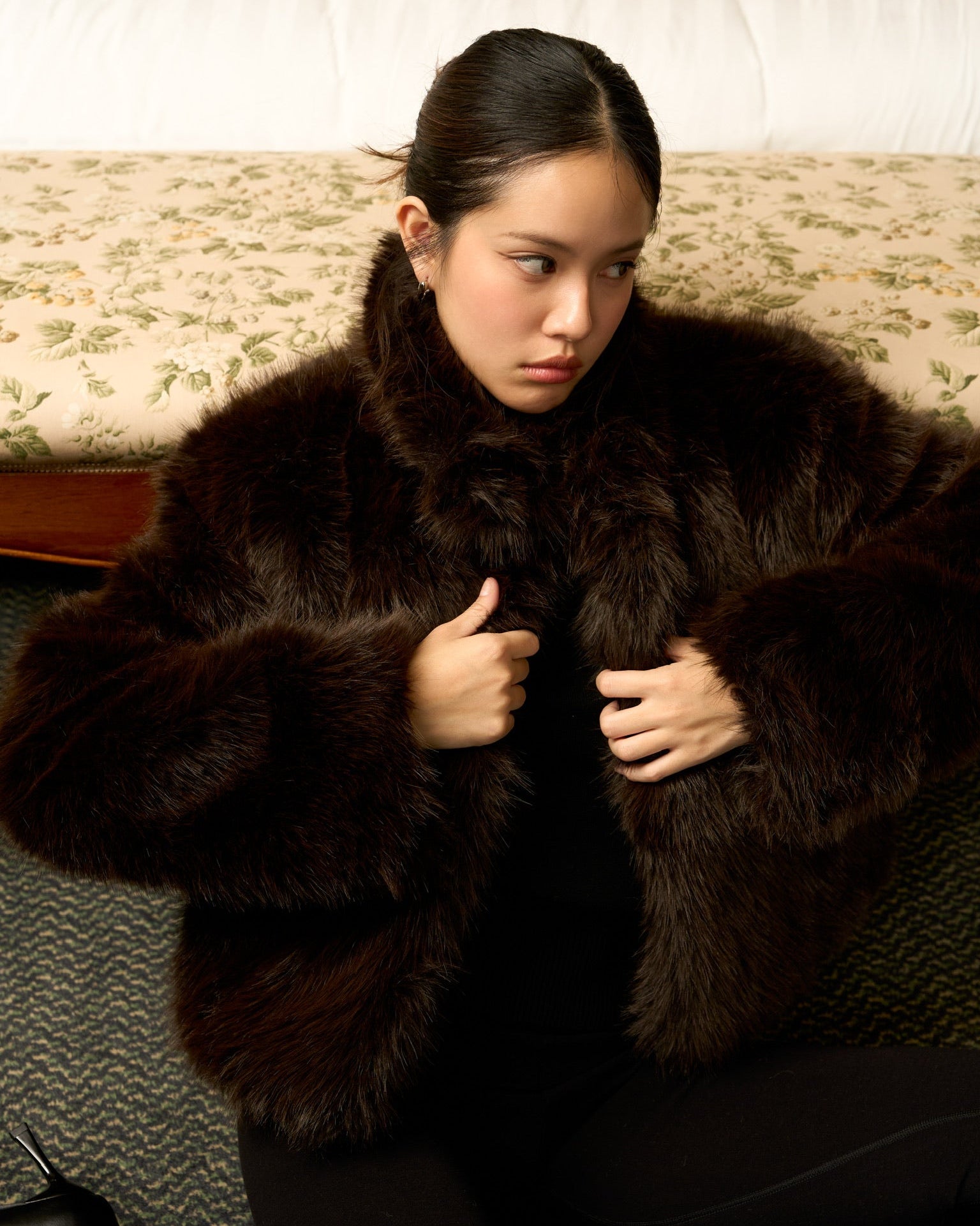 Woman wearing a dark brown fur coat sitting on a patterned couch.