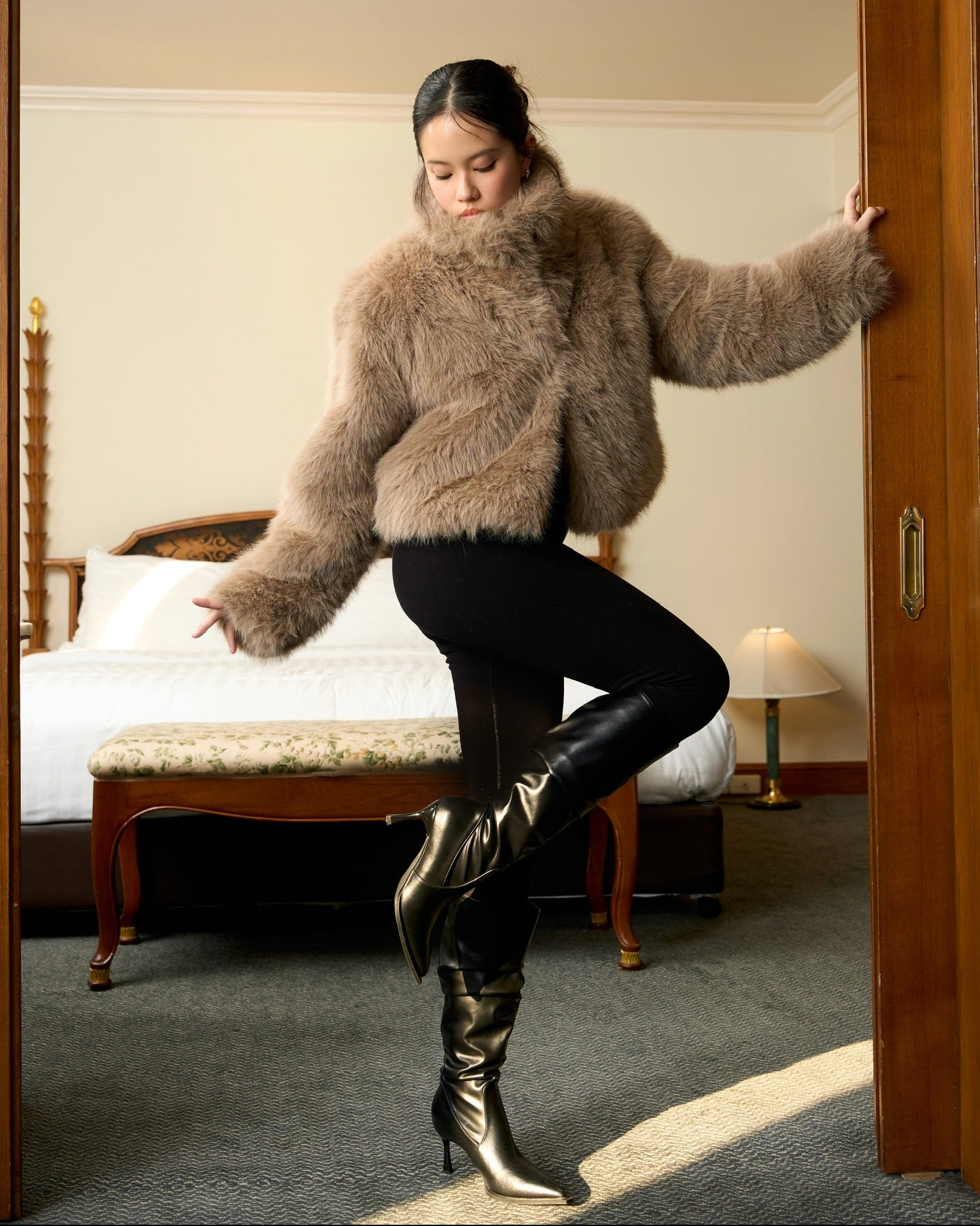 Woman in a fluffy brown jacket and black pants standing in a room with a bed and lamp.