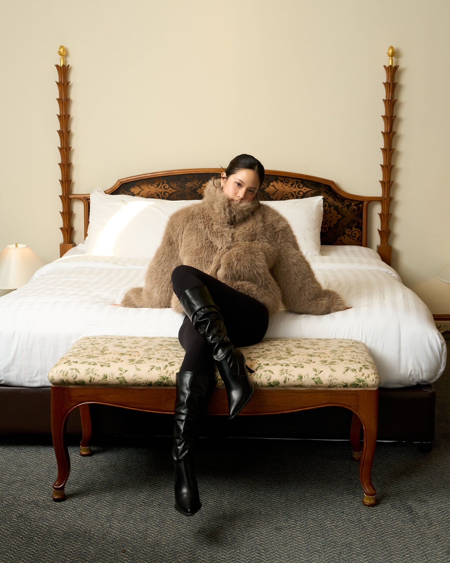 Woman in fur coat and black boots sitting on a bed in a room with wooden furniture.