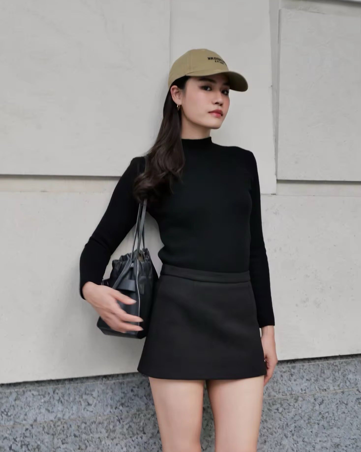 Woman wearing a black outfit with a cap, standing against a light-colored wall.