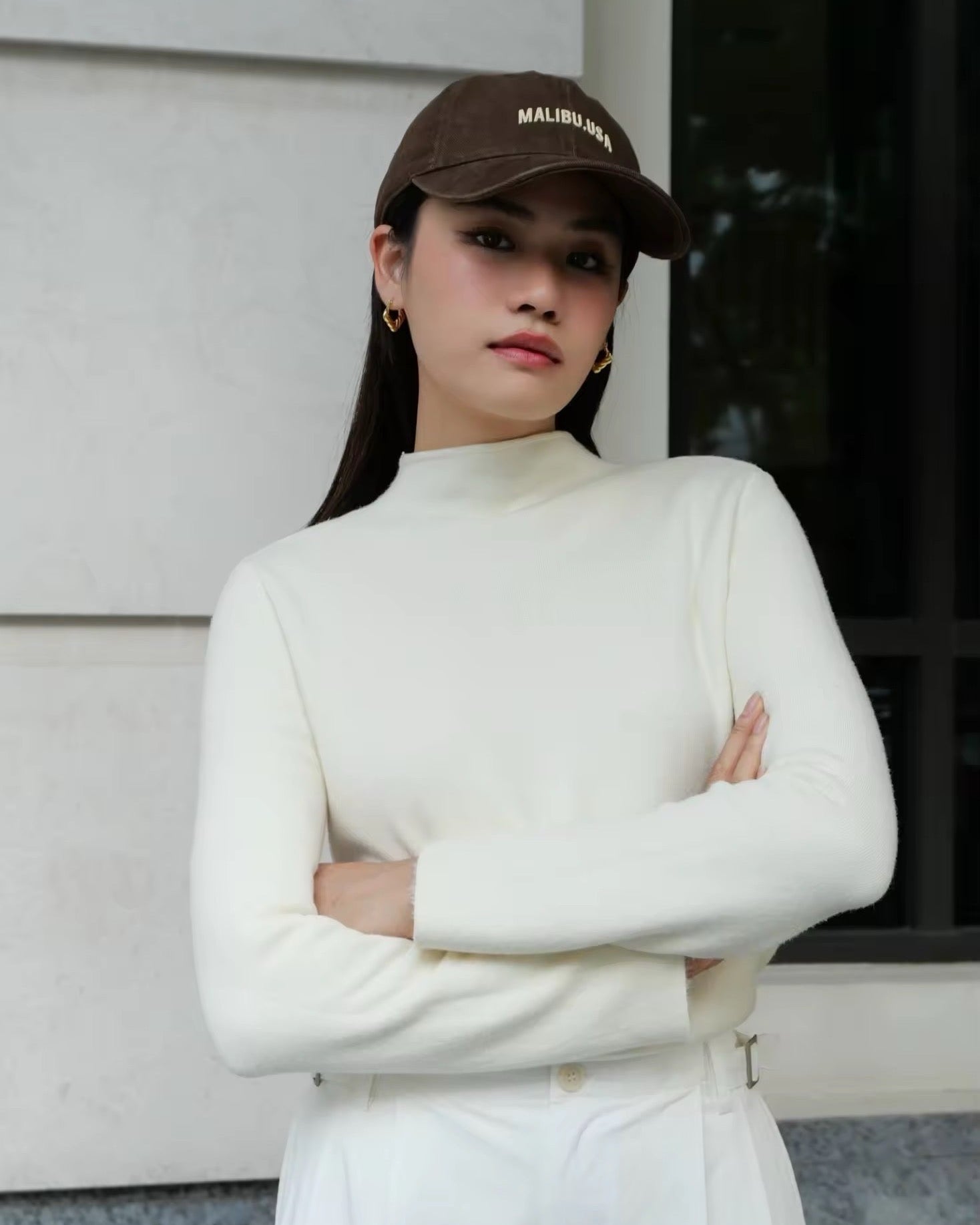 Woman wearing a white turtleneck and cap, standing against a neutral background