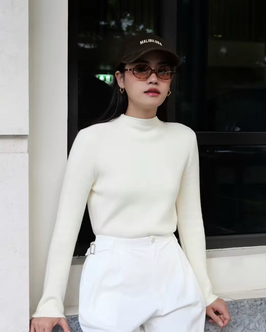 woman wearing a white outfit with a cap and sunglasses, standing in front of a building entrance.