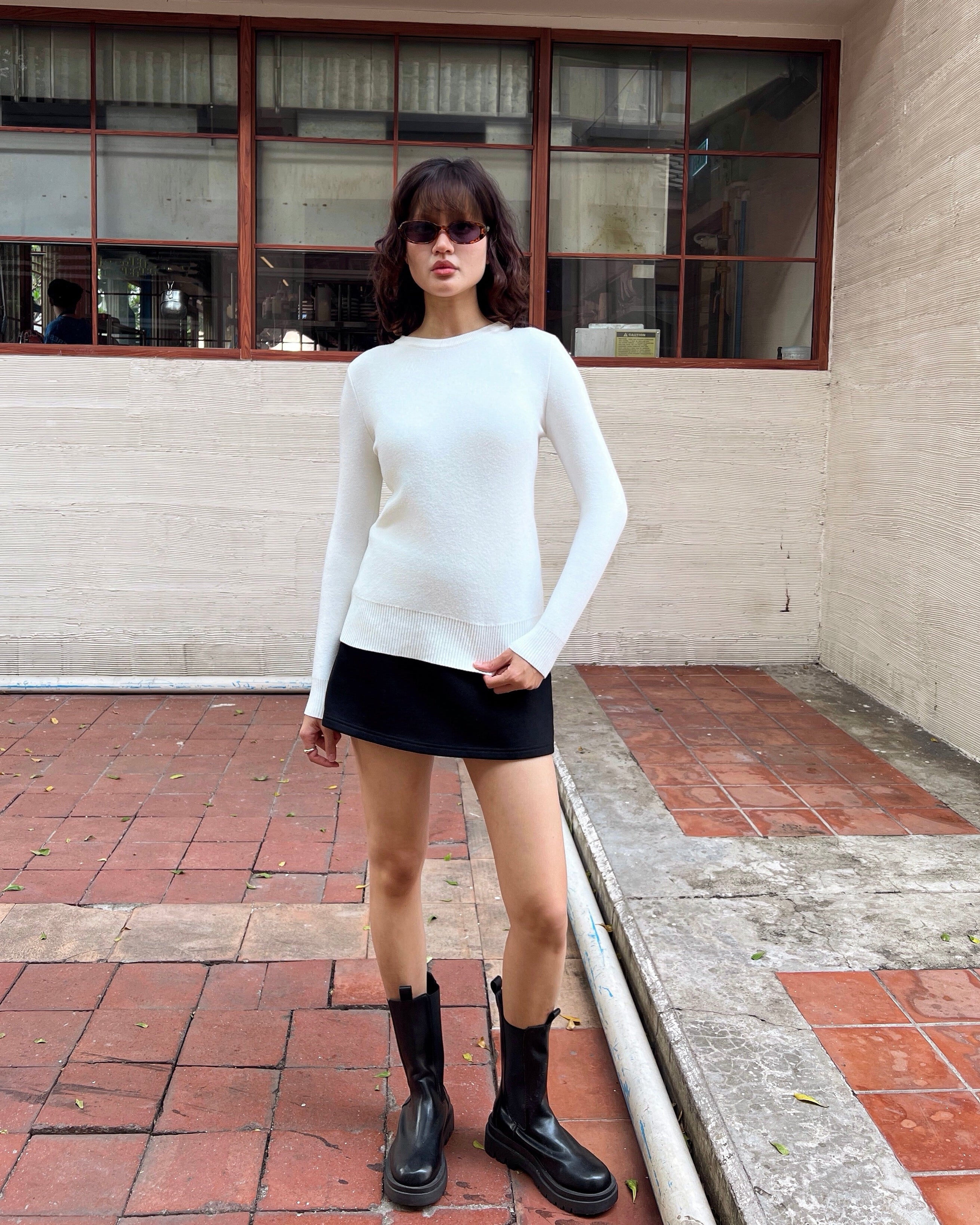 woman wearing a white sweater and black skirt standing in front of a building with large windows.