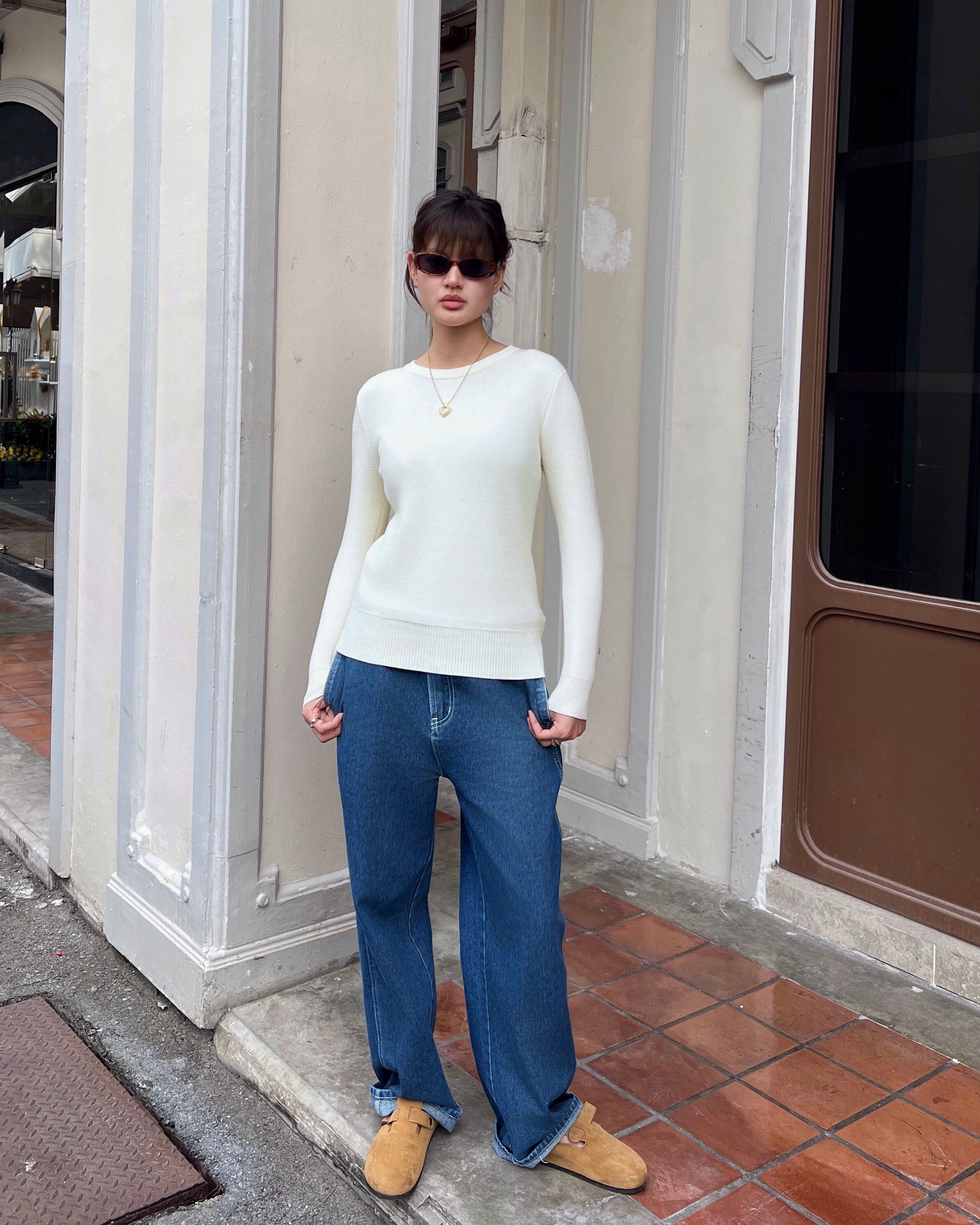 woman wearing a white sweater and blue jeans standing in front of a building entrance.