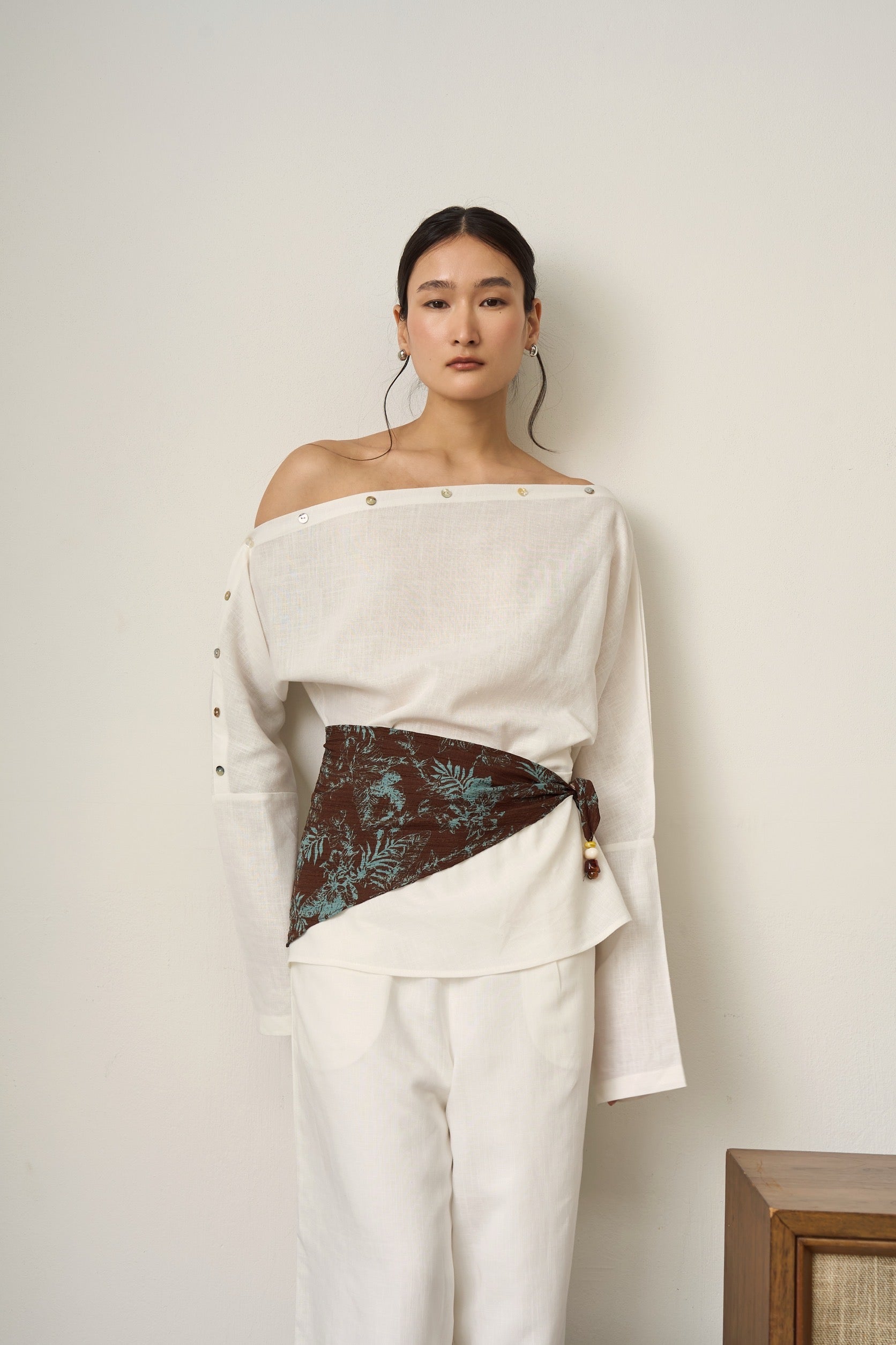 Woman wearing a white off-shoulder linen top with a brown patterned panel and white linen pants against a plain background.