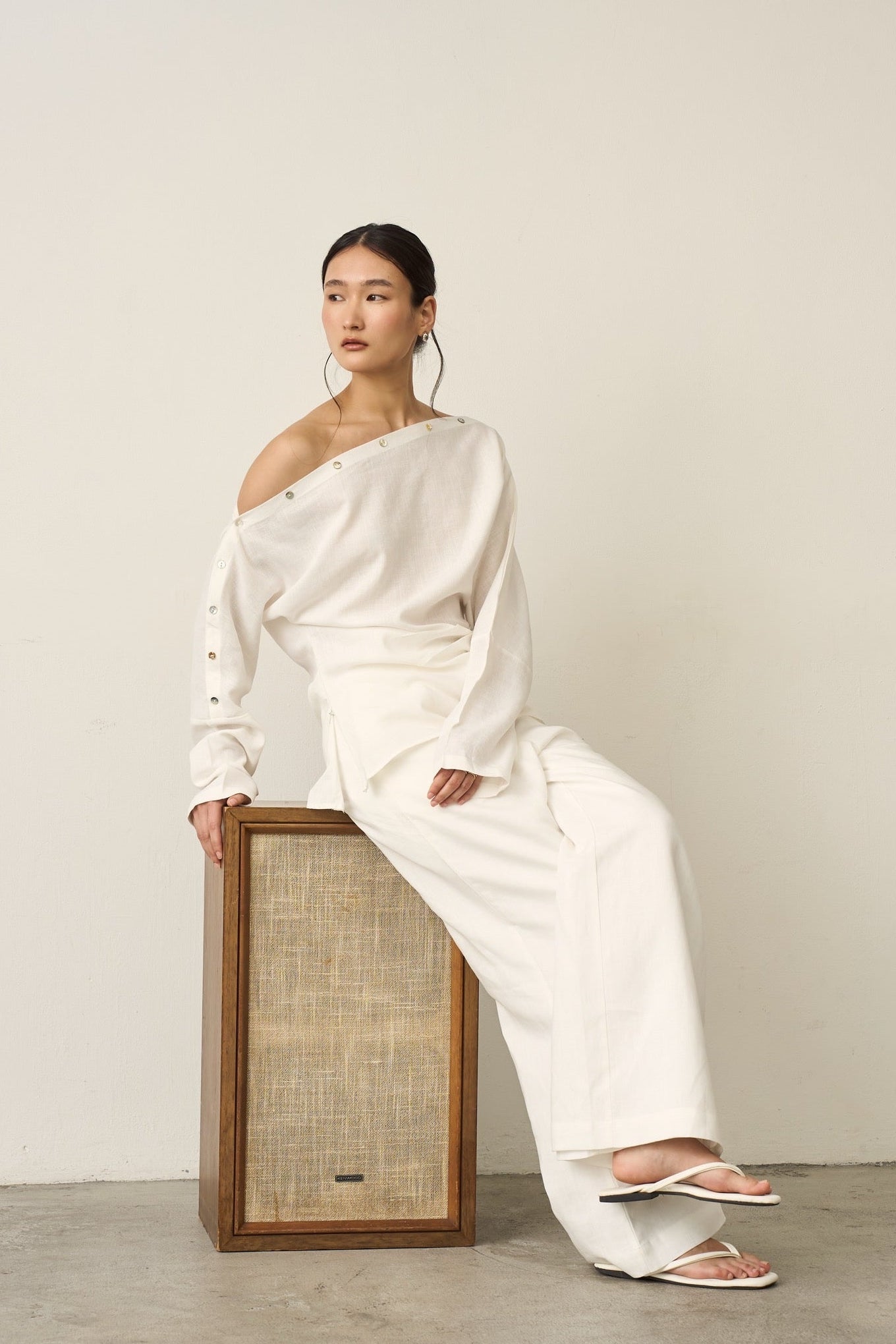 Woman in a white off-shoulder linen shirt and linen pants outfit sitting on a wooden box against a plain background.