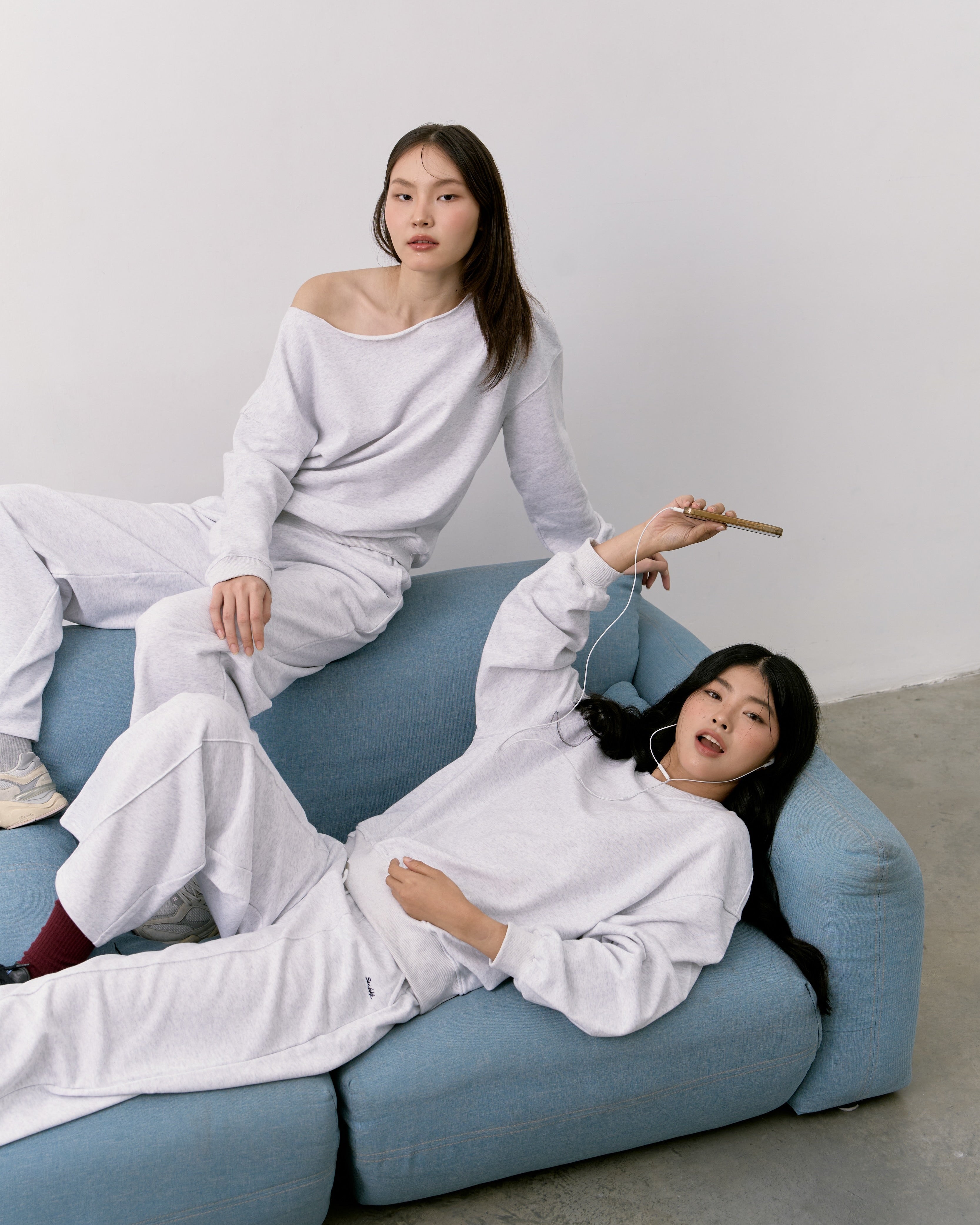 Two women in white outfits sitting on a blue sofa against a plain background