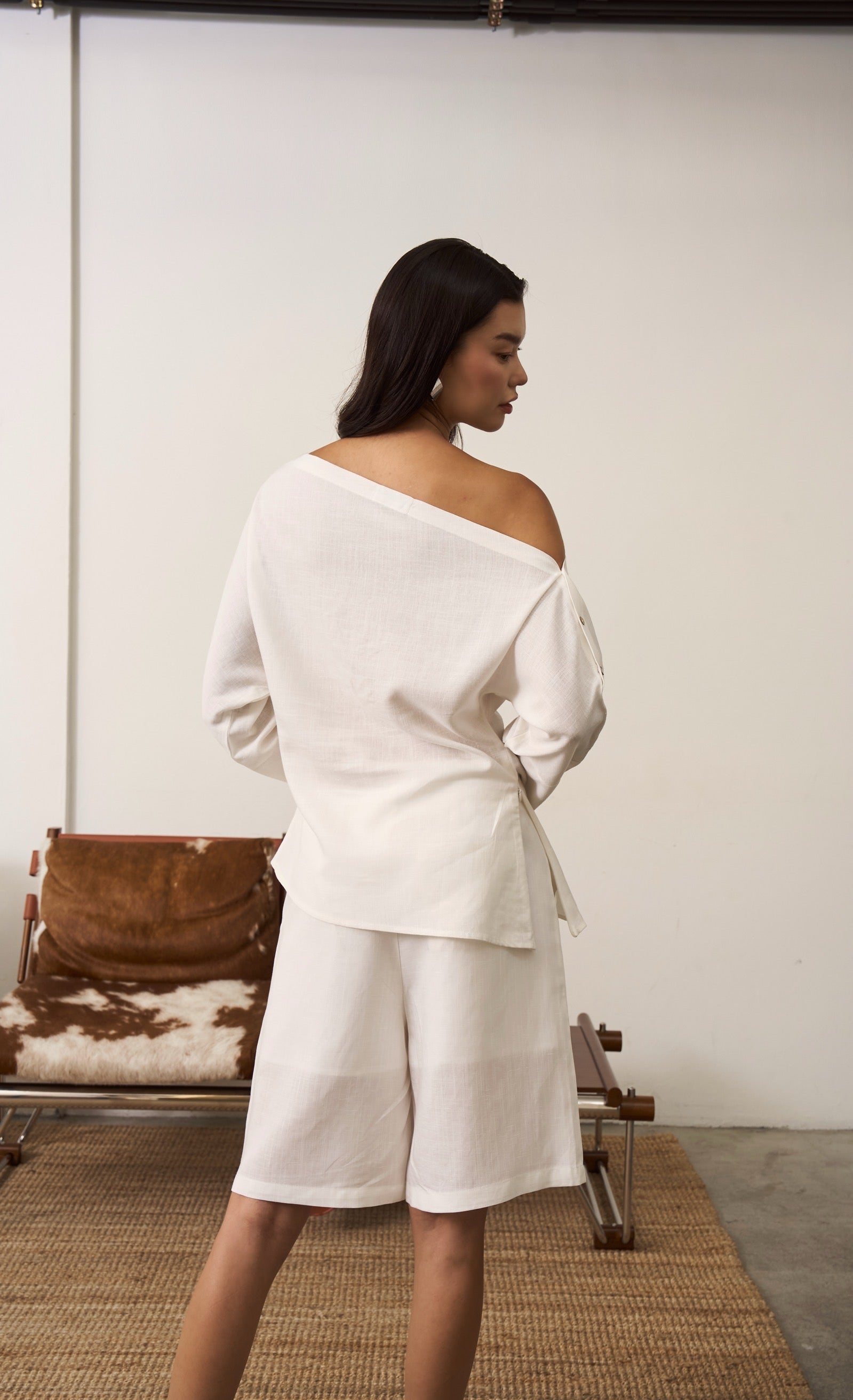 Back of a woman wearing a white off-shoulder linen top and shorts in a minimal indoor setting.