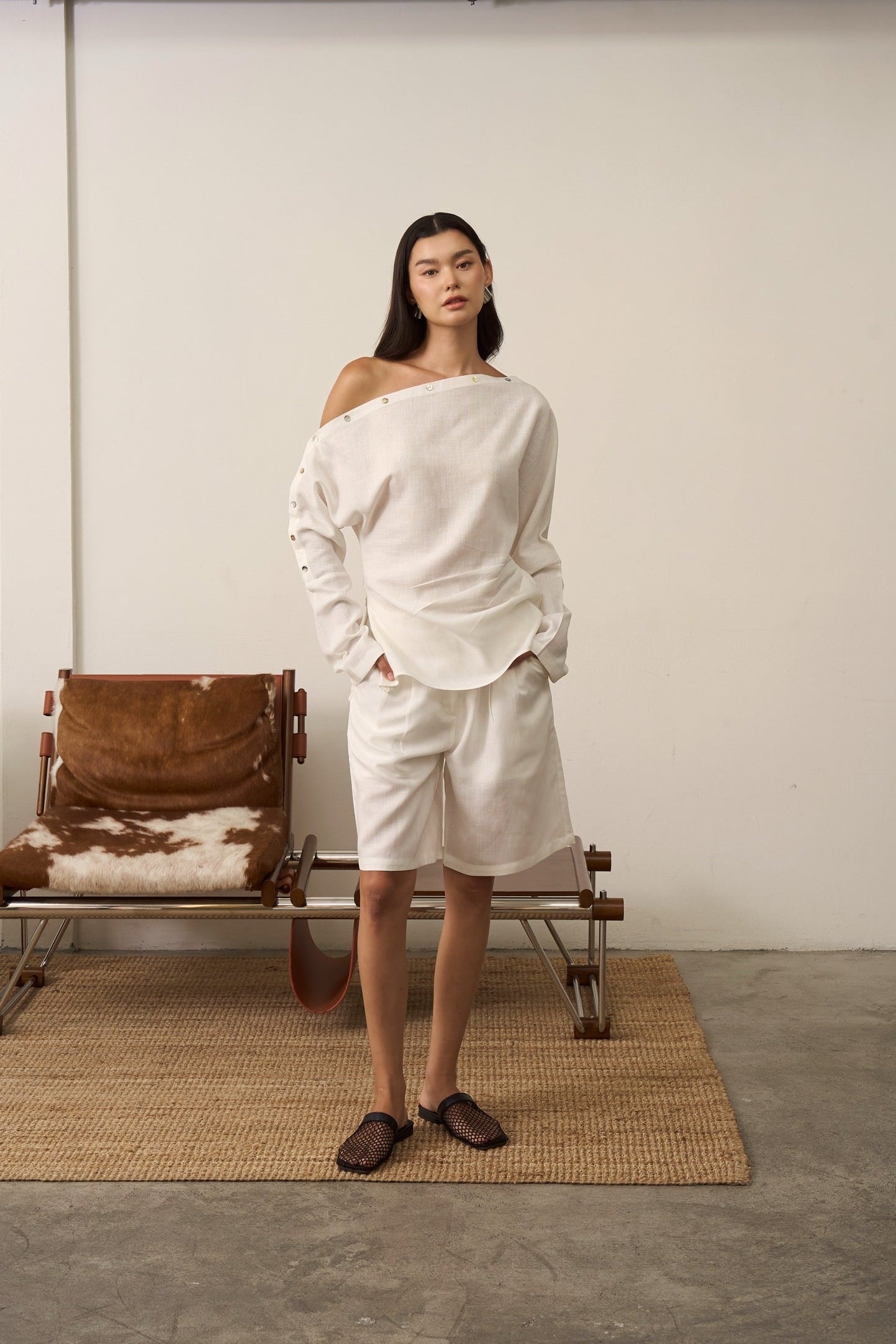 Woman in white linen shirt and linen shorts outfit standing in a minimalistic room with a chair and rug.