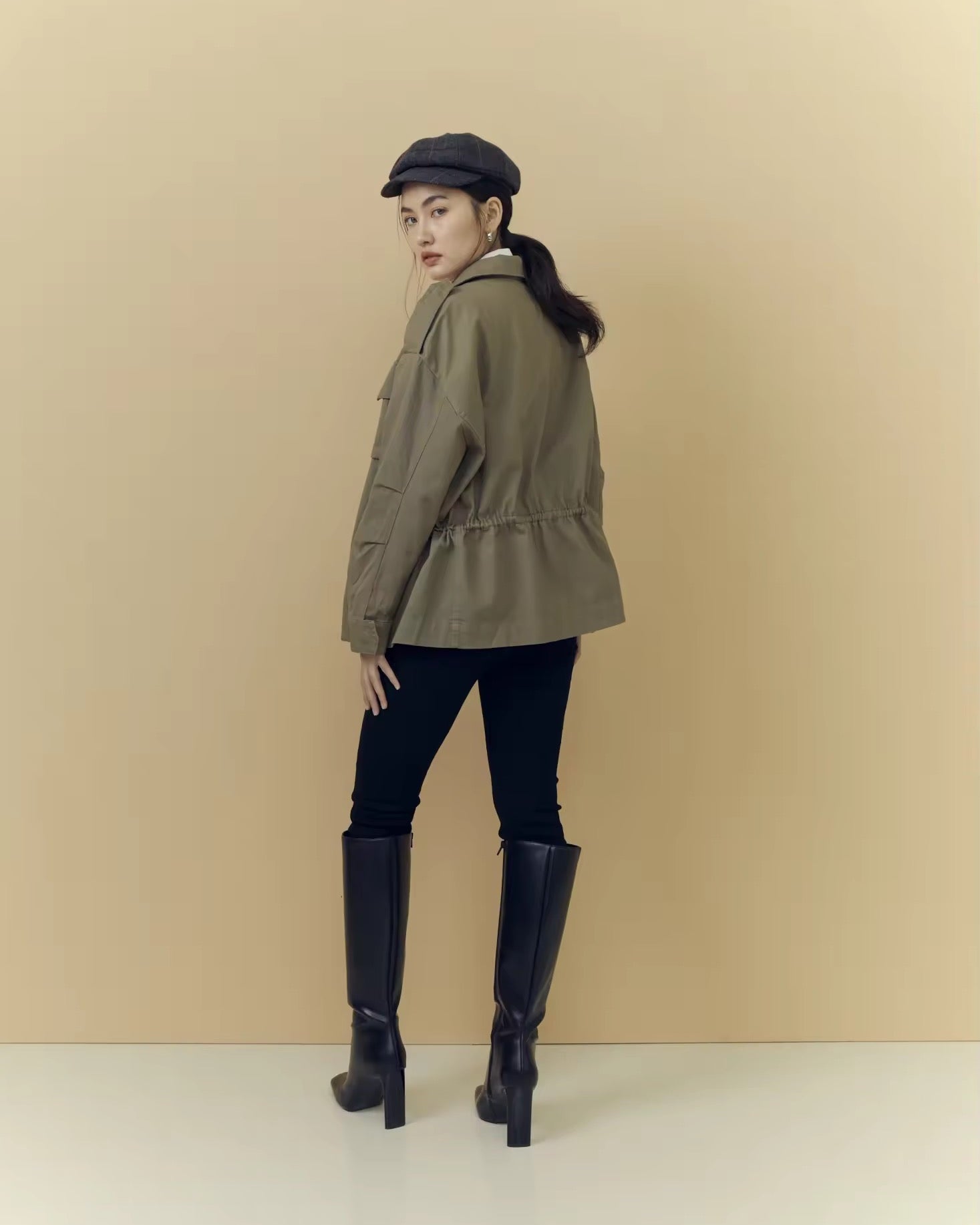 Woman wearing a green jacket, black boots, and a cap against a beige background