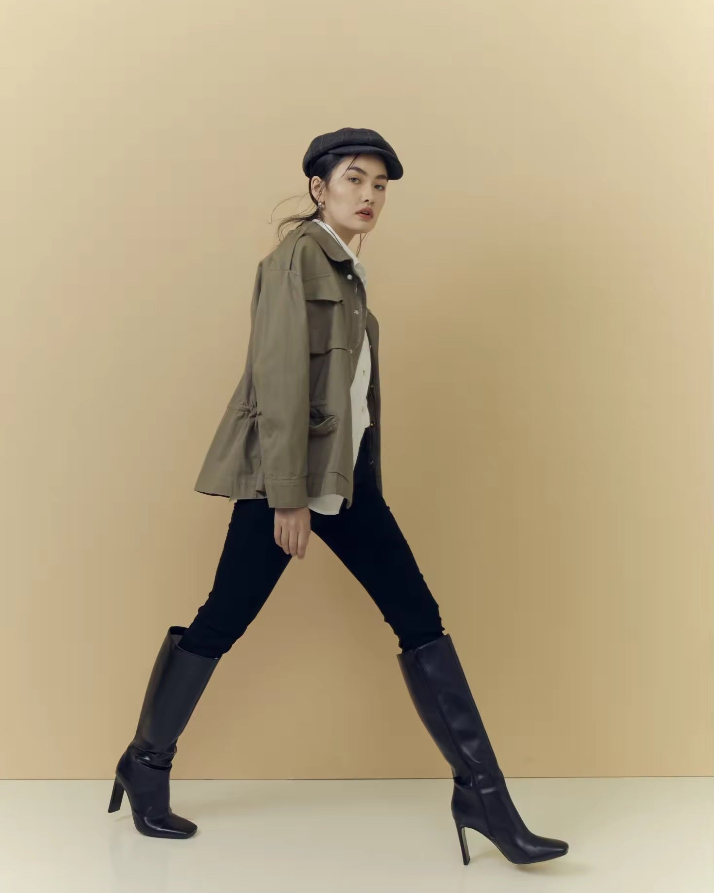 Woman in a green jacket and black boots walking against a beige background