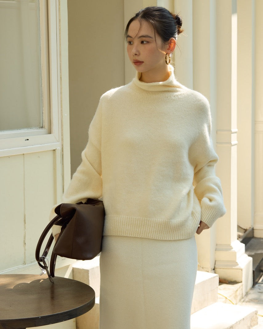 Woman wearing a cream sweater and white pants standing outdoors.